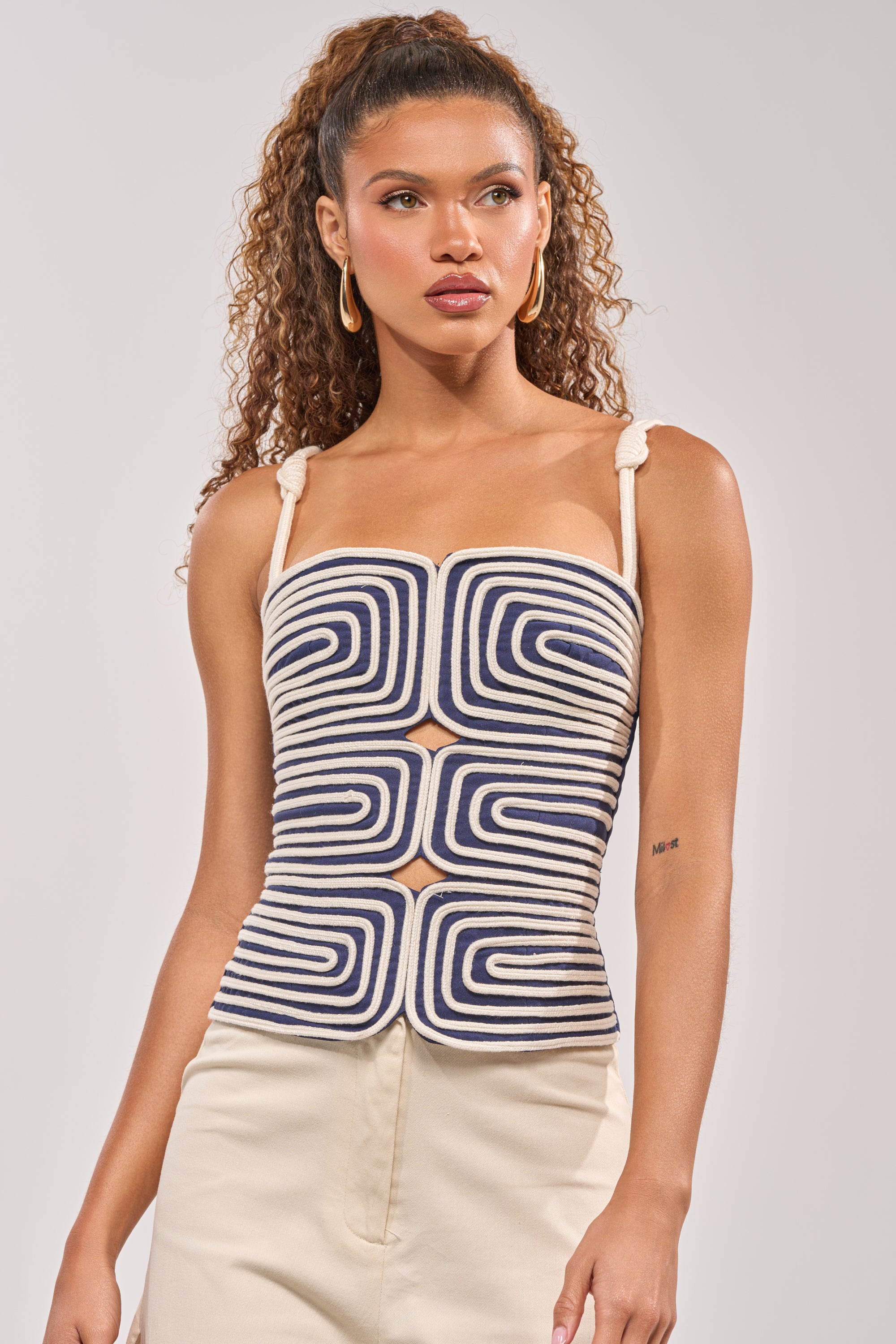 A woman with curly hair wears gold hoop earrings and the DO NOT DISTURB ROPE TANK, a navy blue and white top with sculptural detail and tied straps, paired with light pants against a plain background. 0