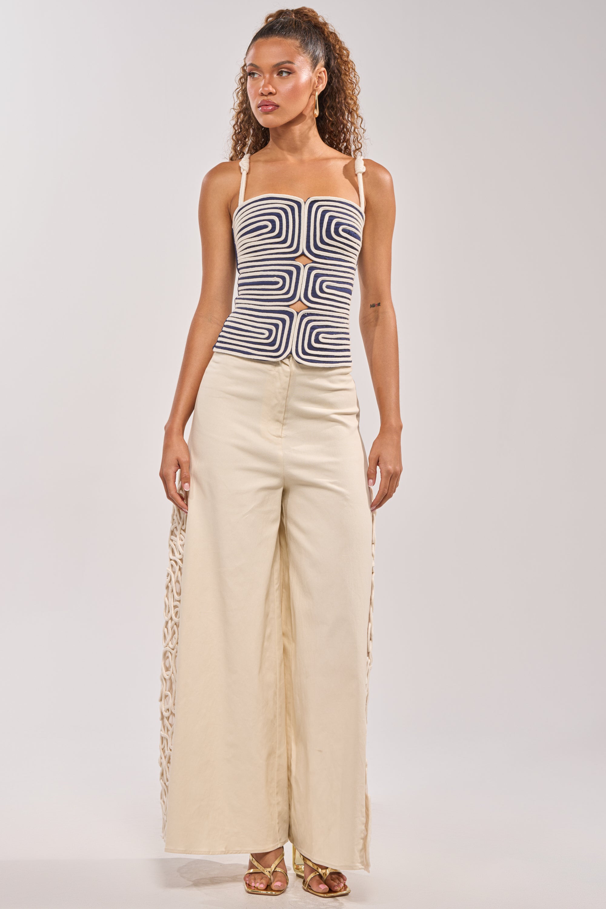 A woman with curly hair wears the DO NOT DISTURB ROPE TANK, a black-and-white geometric tank with shoulder ties, paired with wide-leg beige pants featuring lace side panels and gold high-heeled sandals, against a plain background. 4