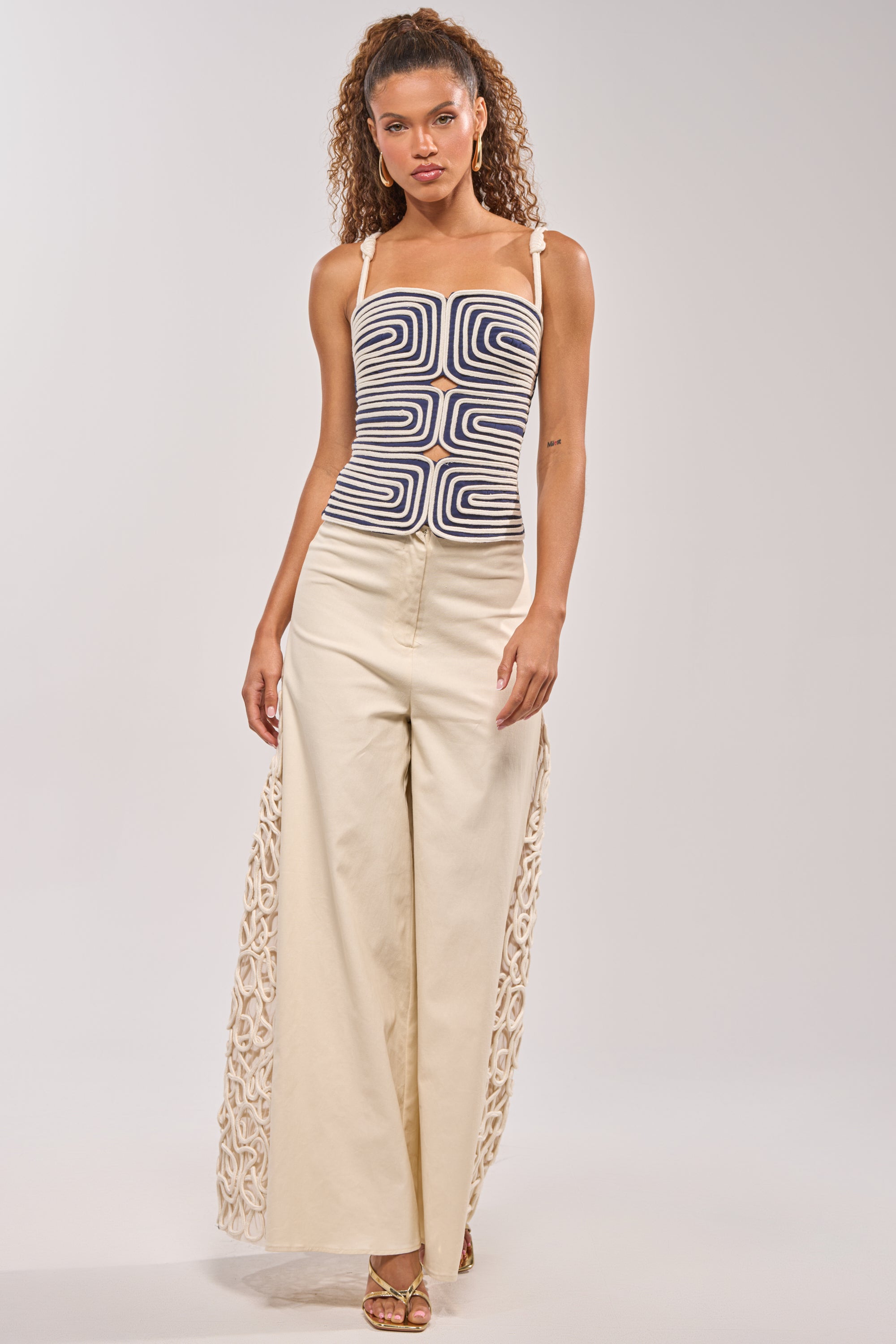 A woman wears the DO NOT DISTURB ROPE TANK, a sleeveless top with a bold black and white geometric pattern, paired with high-waisted wide-leg cream pants featuring sculptural side details. She completes the look with heeled sandals and effortless curls. 5