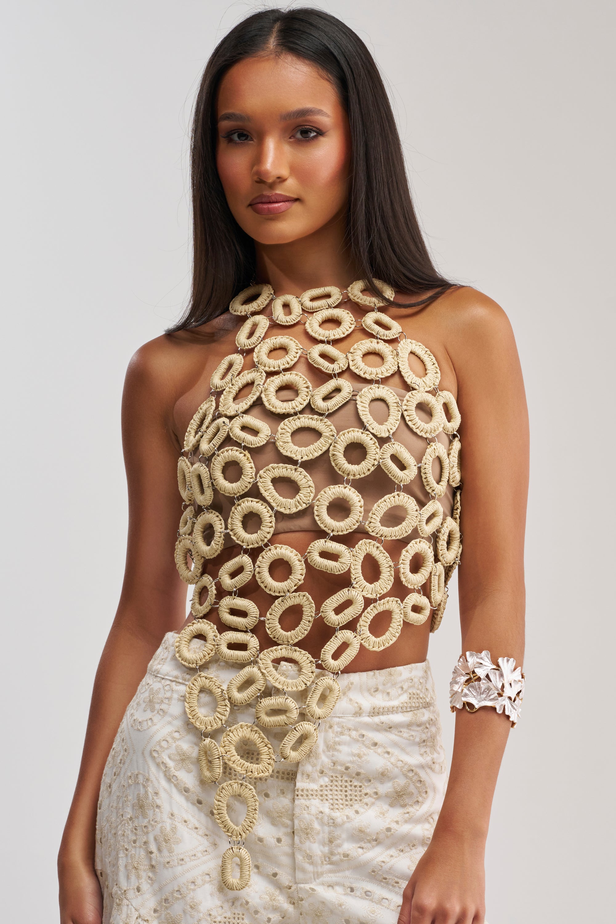 A woman with long dark hair wears the BEACH VIBES CHAIN TOP, featuring large crocheted loops, paired with white patterned pants and a chunky coral-shaped bracelet. She stands against a plain light background.