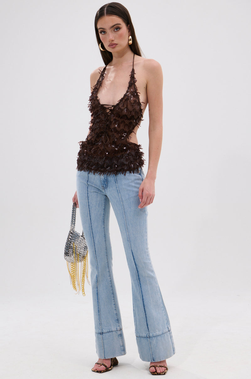 A woman poses against a plain background wearing the SEQUIN FRINGE QUEEN HALTER NECK BLOUSE in brown, paired with light blue flared jeans, high-heeled sandals, a metallic beaded purse, large earrings, and long straight hair. 4