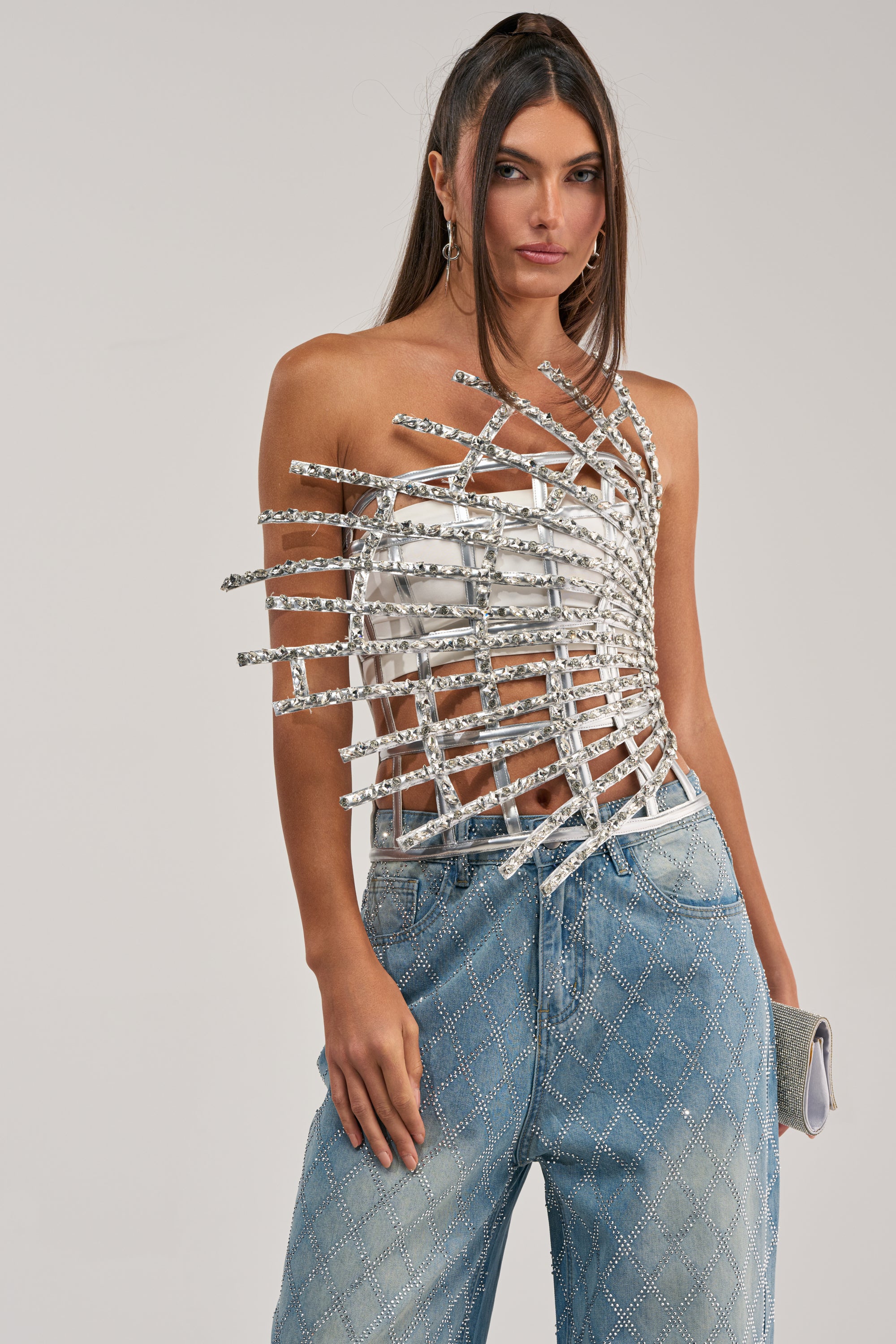 A woman wears the MADE YA LOOK RHINESTONE CAGE CORSET—a unique silver cage corset top—over a strapless white bandeau, paired with light blue embellished jeans and holding a small silver clutch.