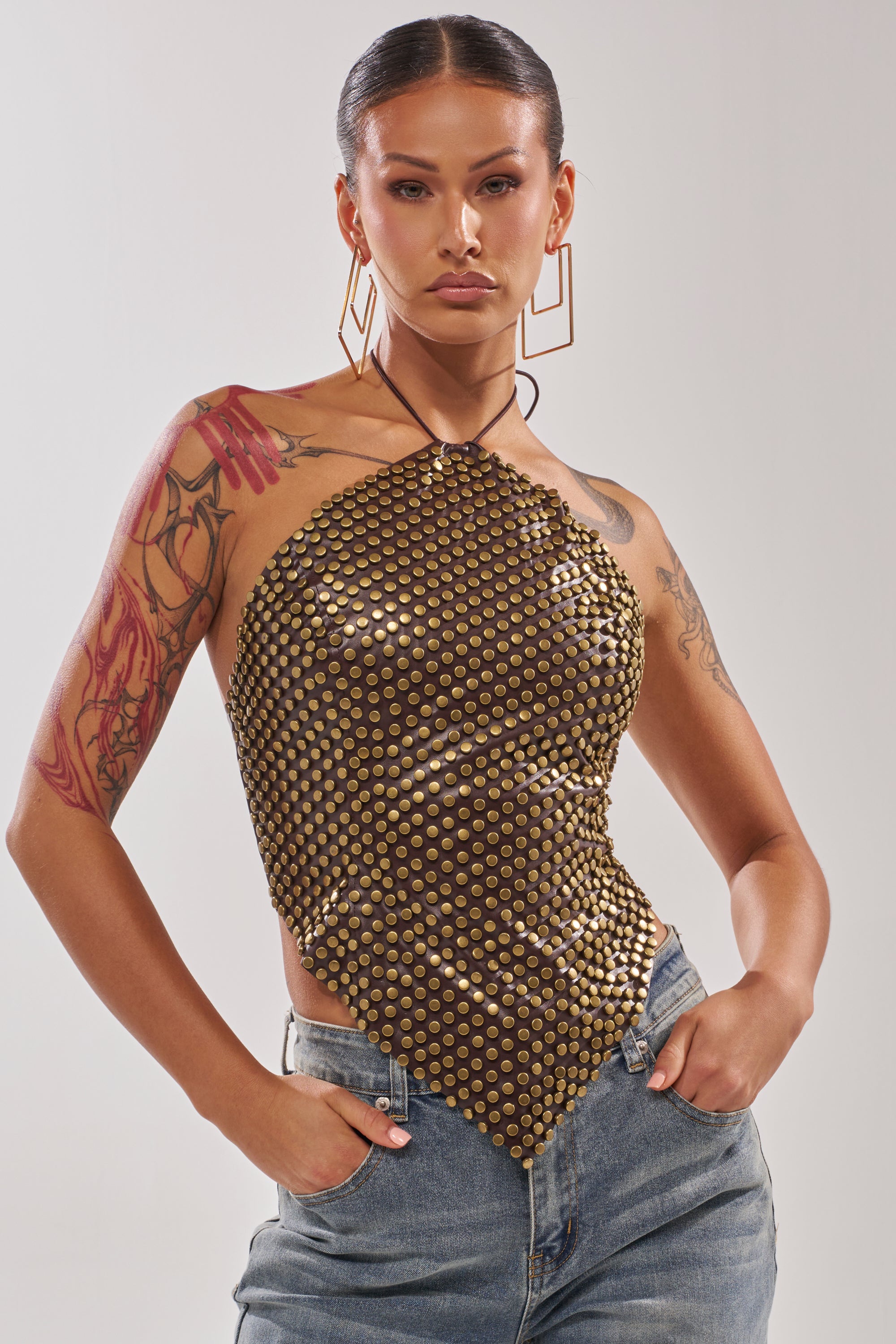 A person with slicked-back hair wears the GET FUNKY BEADED TOP IN BROWN and blue jeans, posing with hands in pockets. They have visible arm tattoos and large geometric earrings, standing against a plain background. 0