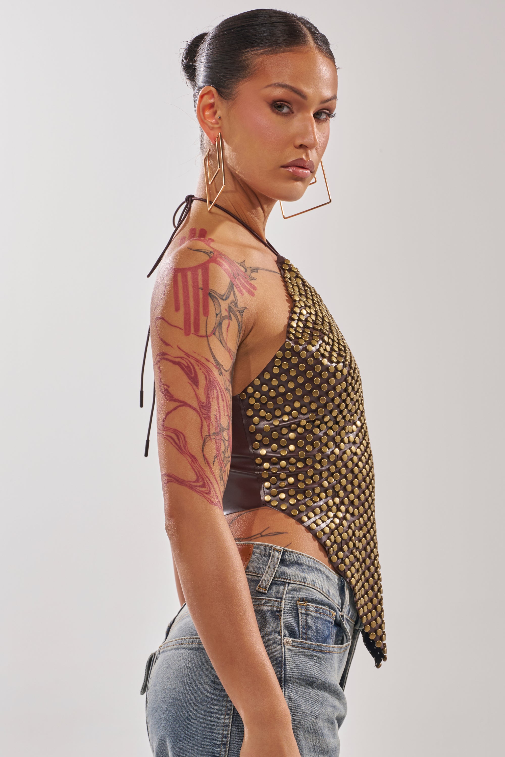 A woman with dark hair in a bun models the GET FUNKY BEADED TOP IN BROWN with blue jeans, large geometric earrings, and visible arm tattoos, confidently glancing over her shoulder against a plain background. 2