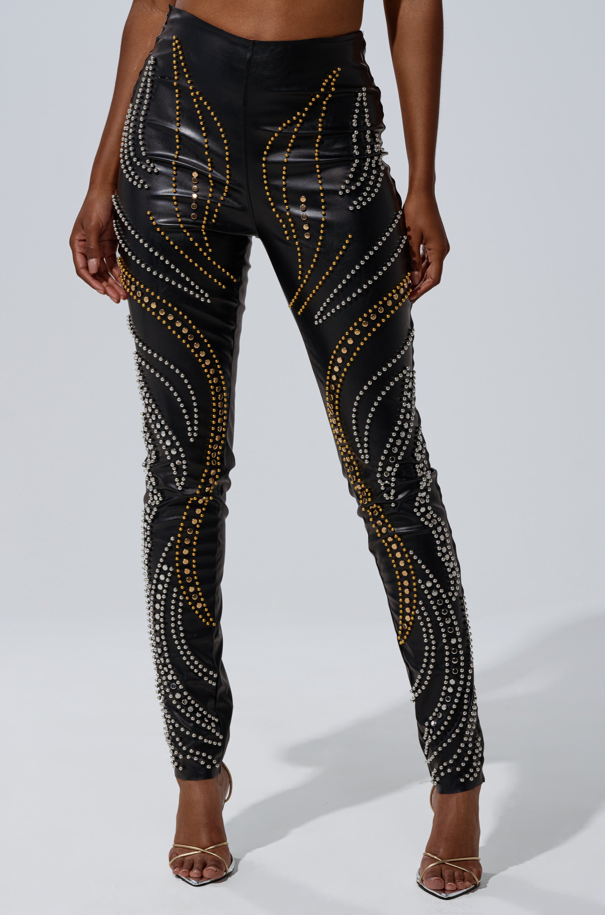 A person models the LETS GET FUNKY BEADED LEGGING—high-waisted faux leather leggings with gold and silver beaded swirl patterns—paired with open-toed heels, shown from the waist down.