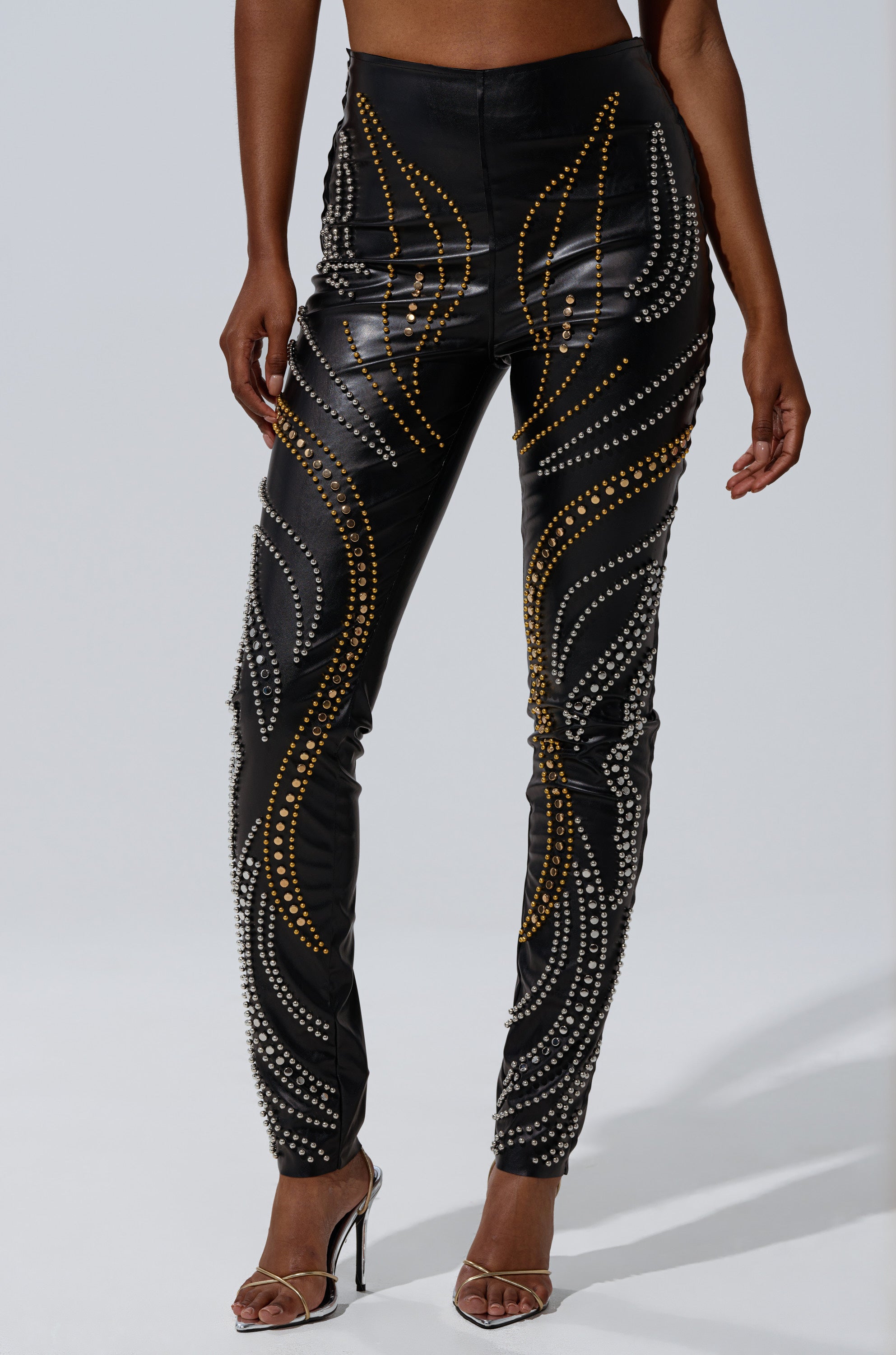A person wears LETS GET FUNKY BEADED LEGGING, featuring high-waisted black fabric embellished with swirling gold and silver studs, paired with silver high-heeled sandals. The upper body is not visible.