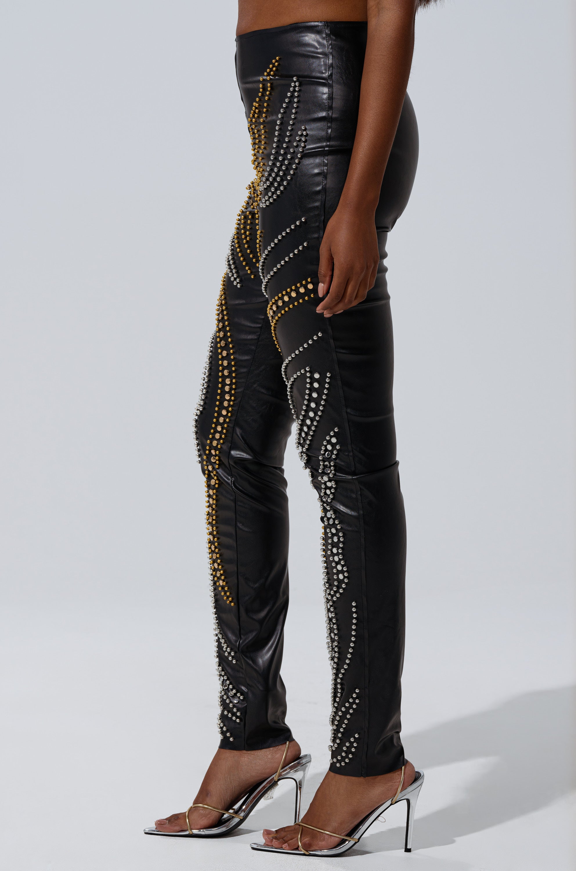 A person wearing LETS GET FUNKY BEADED LEGGING, black faux leather leggings adorned with gold and silver beaded swirl patterns, paired with silver high-heeled sandals, stands against a plain white background.
