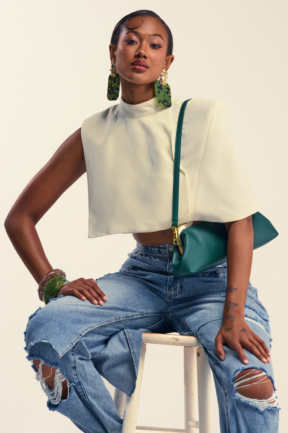 A woman, exuding confidence with a side-swept hairstyle, sits on a stool wearing the DRESSED TO IMPRESS BLOUSE in cream paired with ripped blue jeans. She accessorizes her look with large green earrings, a matching green bangle, and holds a green purse.