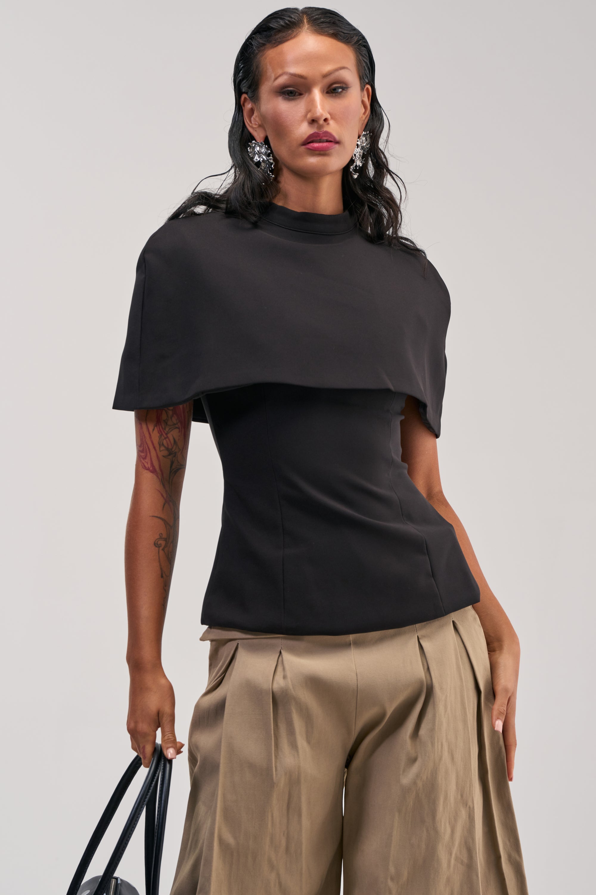 A woman poses in the TIME TO GET SERIOUS BLOUSE, featuring a dramatic black cape-like overlay, paired with wide tan pleated pants and statement earrings. She holds a black bag and wears her dark hair in a sleek, wet-look style against a plain light background.