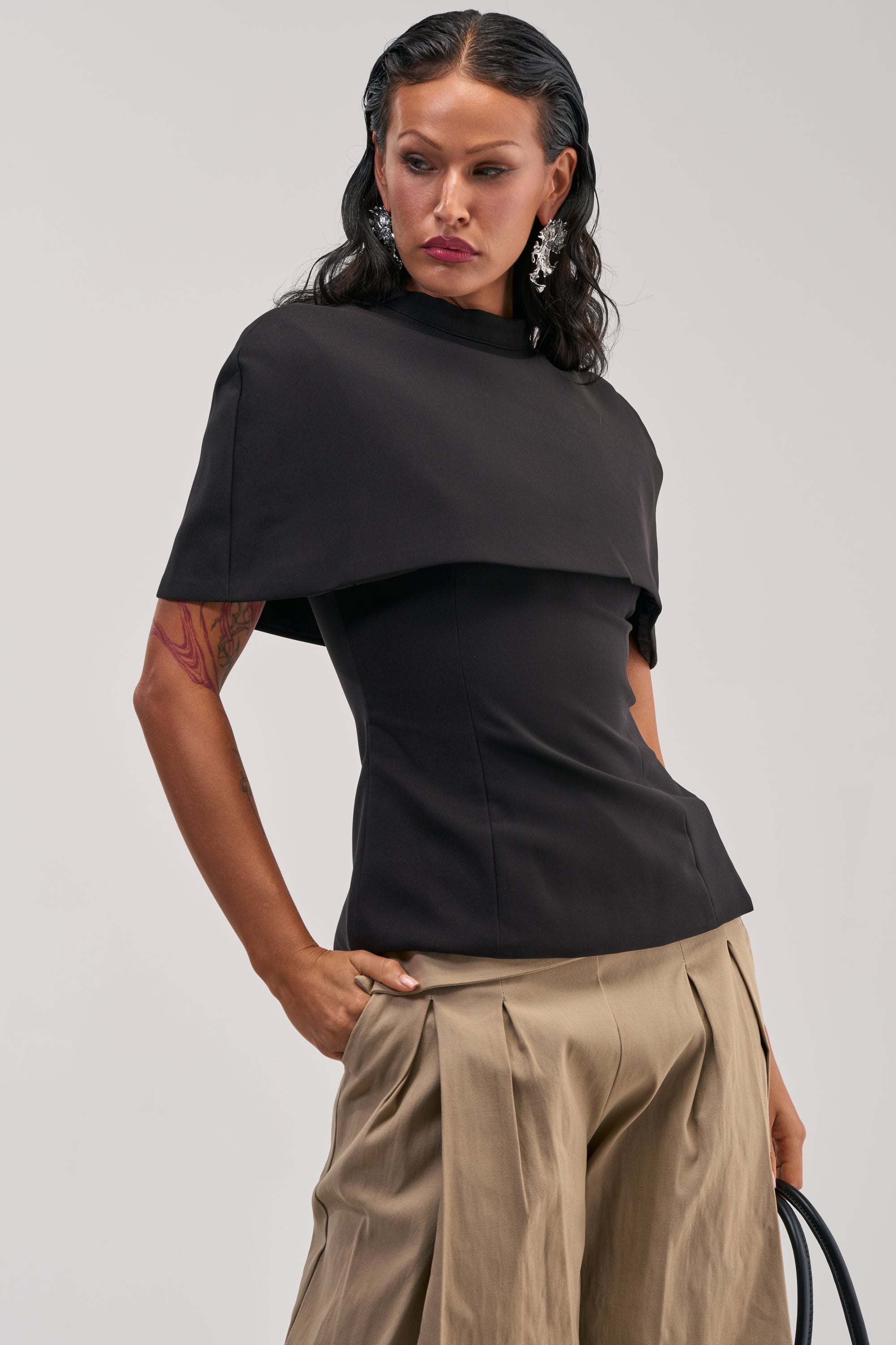 Wearing the TIME TO GET SERIOUS BLOUSE, a black short-sleeved top with an asymmetrical overlay, this person pairs it with beige pleated pants and a black bag, accessorizing with silver earrings and styling their wavy dark hair.