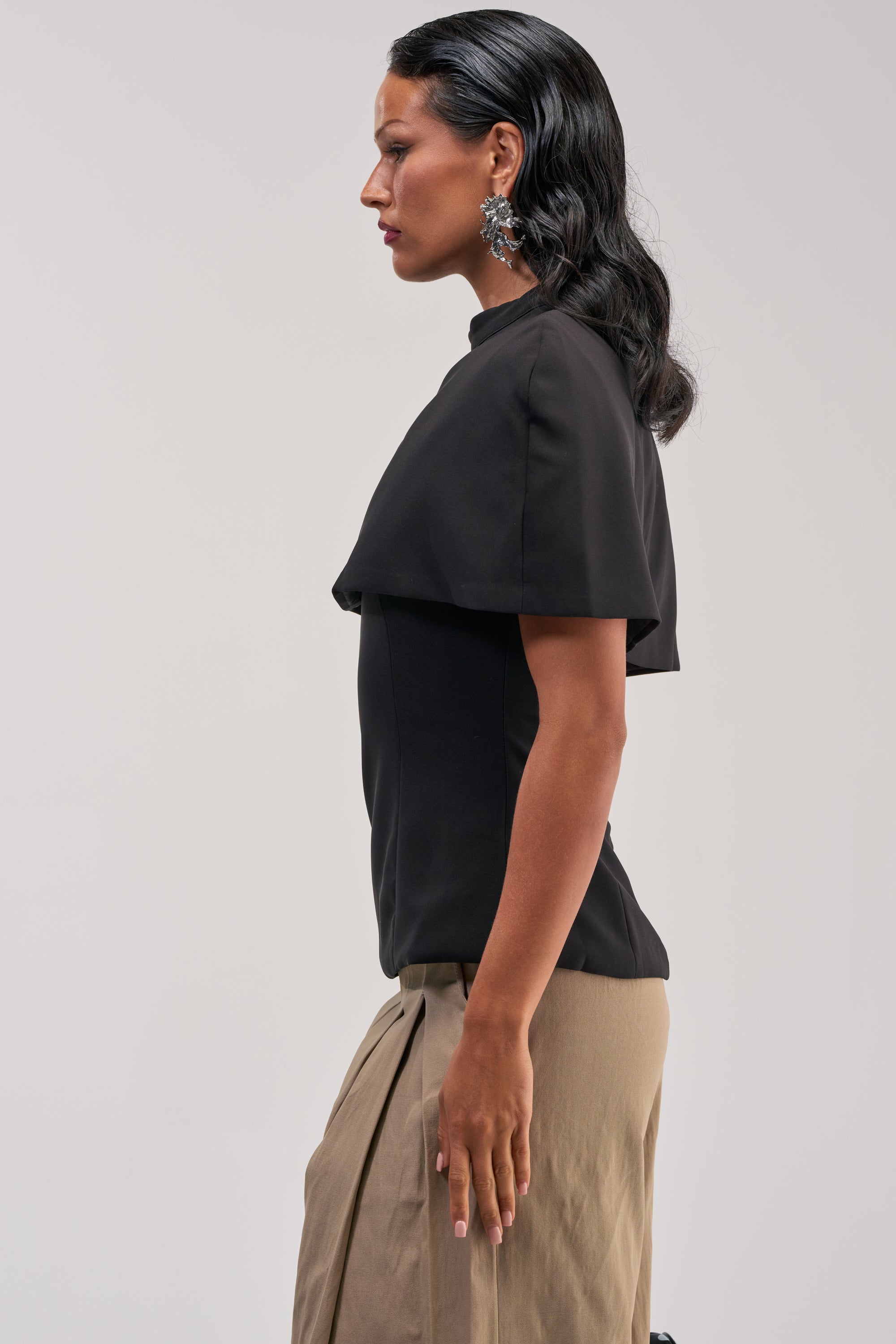 A woman with long dark hair stands in profile against a plain, light background, wearing the TIME TO GET SERIOUS BLOUSE—a black capelet blouse—paired with a beige pleated skirt and large floral earrings.