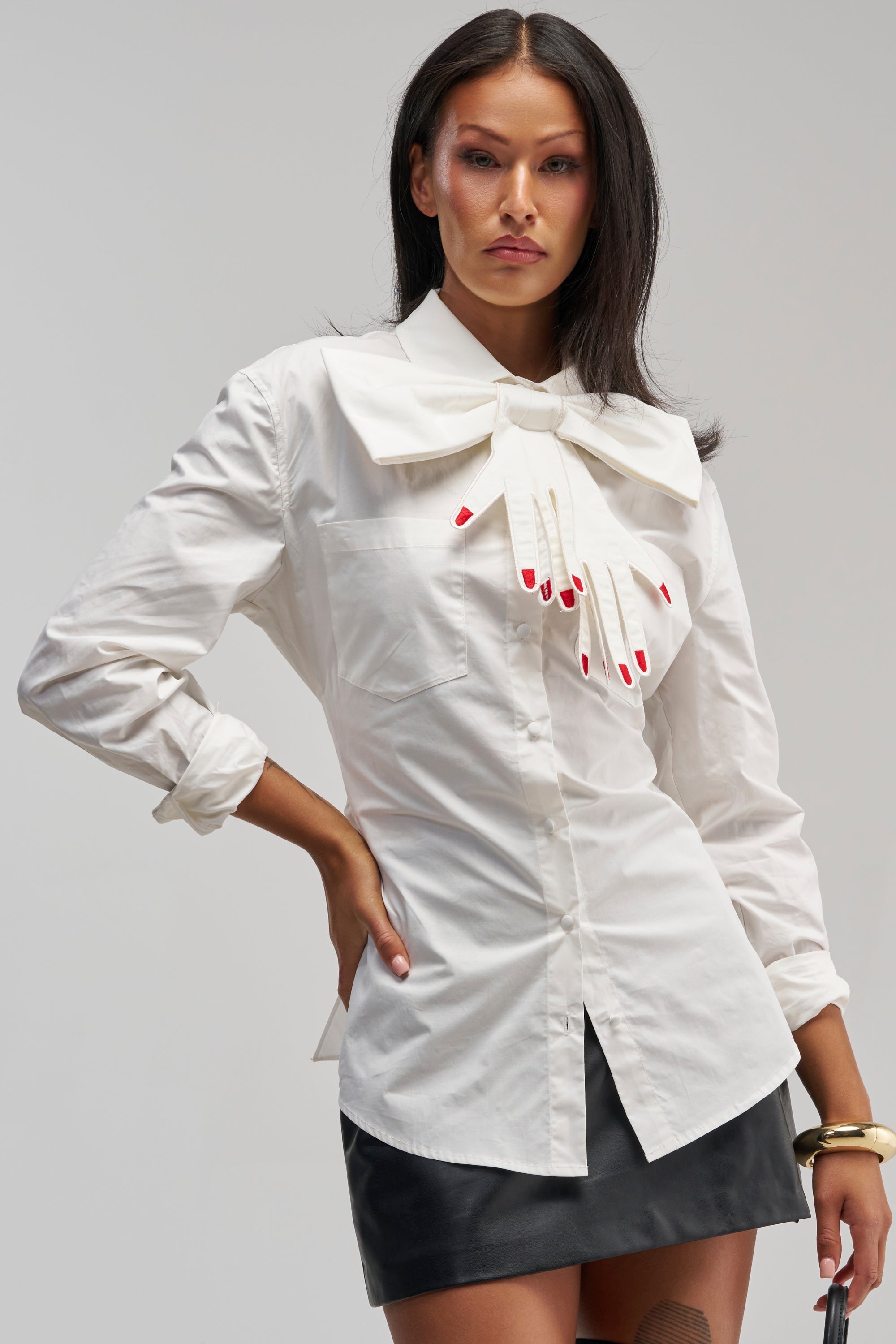 A woman confidently poses against a plain background, channeling modern business casual elegance, in the HANDS OFF POPLIN BLOUSE—a crisp white design featuring an oversized bow shaped like a hand with painted nails—styled with a black mini skirt for the ultimate office siren look. One hand rests on her hip, embodying bold sophistication.