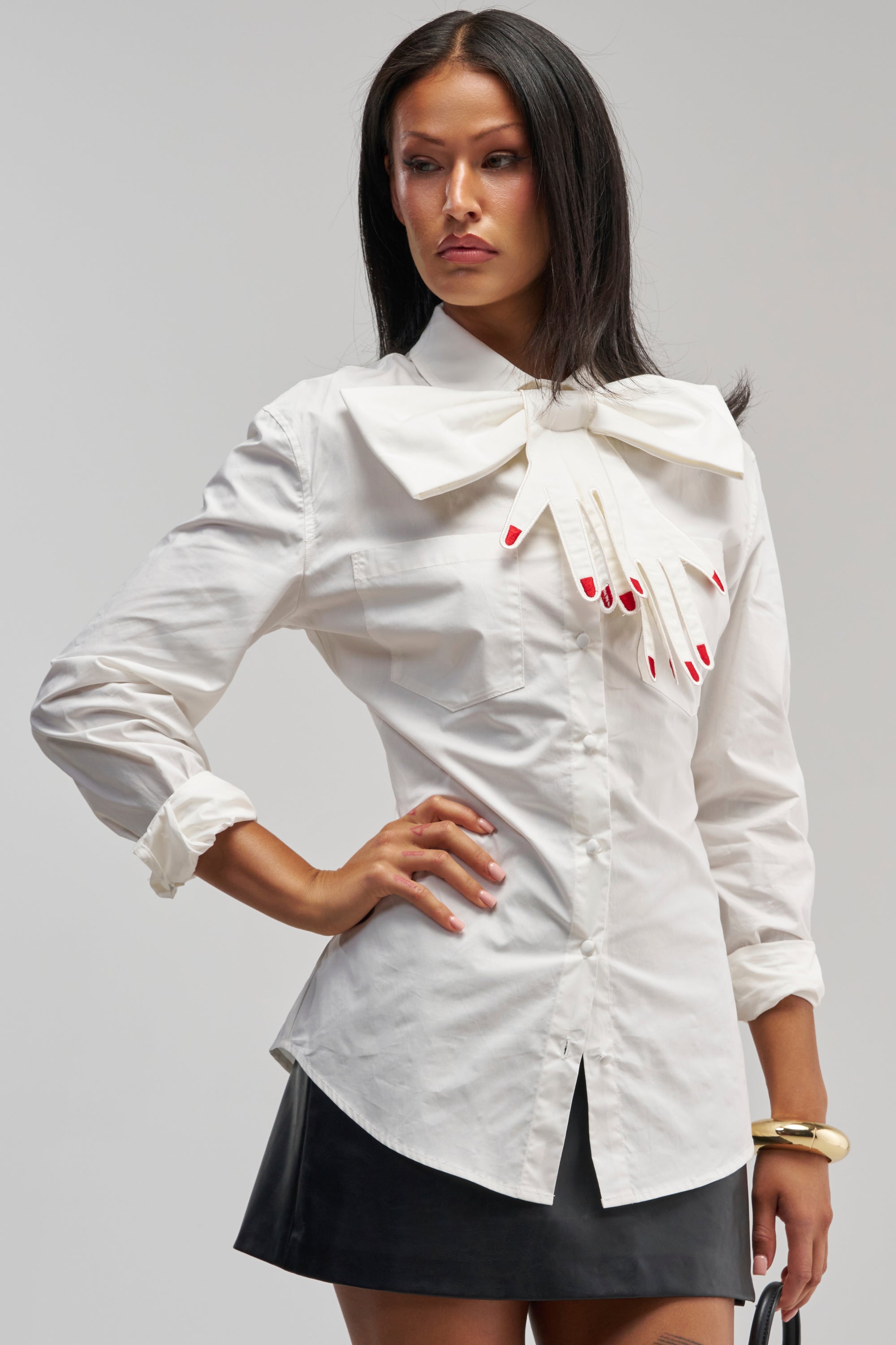 A woman with straight dark hair and a serious expression poses with one hand on her hip, styled in the HANDS OFF POPLIN BLOUSE—a crisp white shirt accented by an oversized collar bow and a whimsical gloved hands motif with red fingernails, delivering an effortlessly chic office siren twist on business casual.