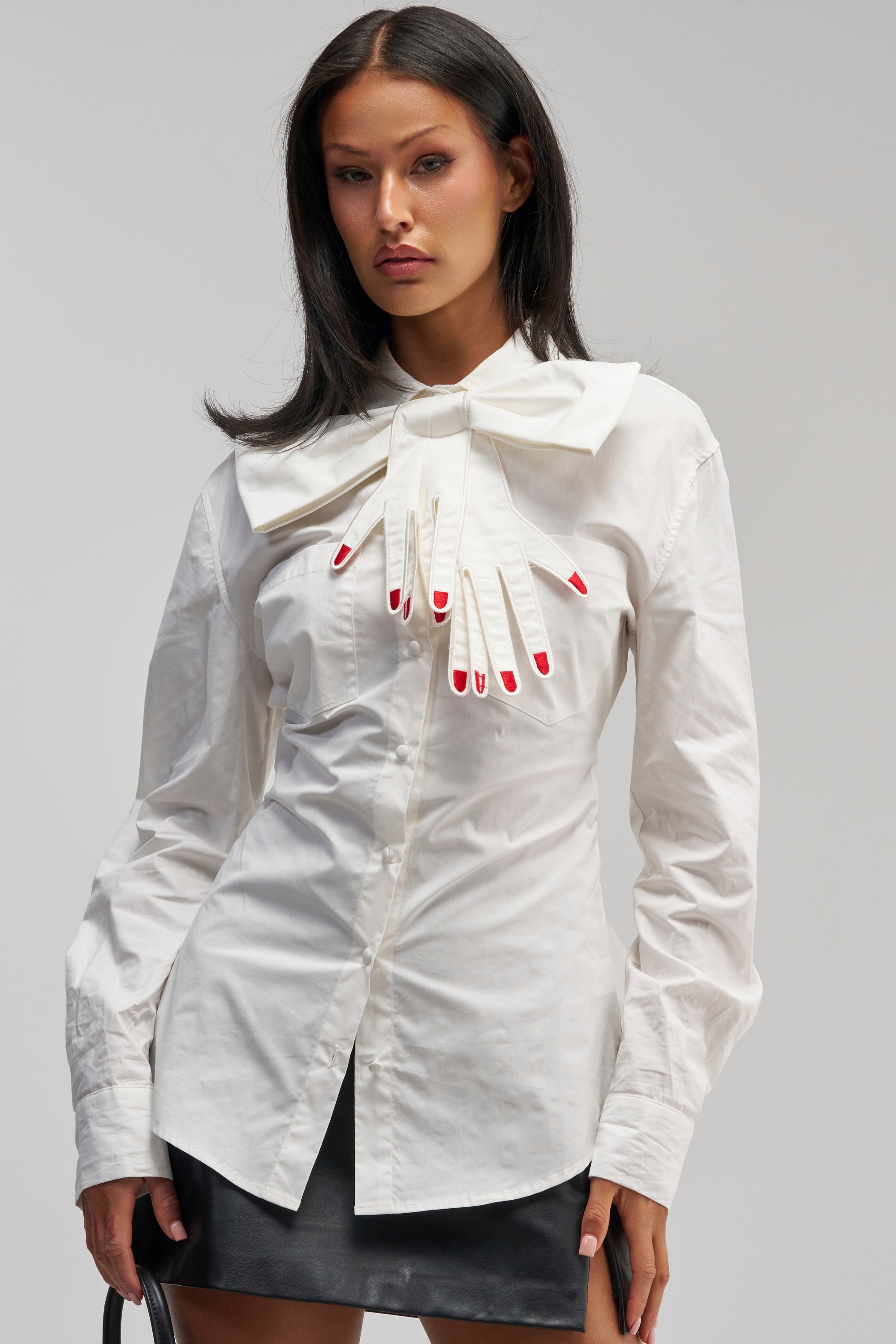 Wearing the HANDS OFF POPLIN BLOUSE—a crisp white top accented with a statement bow and a unique red-nailed glove appliqué—paired effortlessly with a black skirt, she exemplifies chic office siren style against a minimalist backdrop.