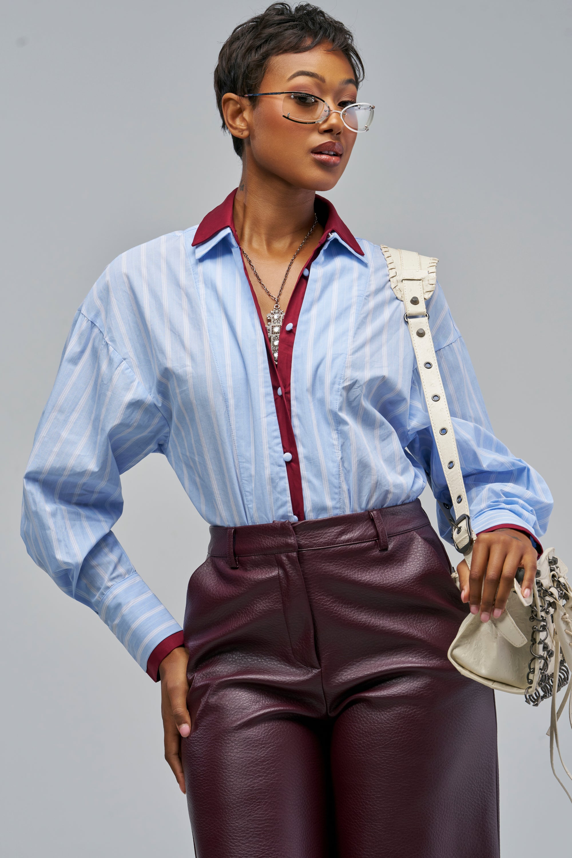 A person wearing a DOUBLE COLLAR TWO-TONE BODYSUIT in blue and white pinstripe layered over a maroon collared shirt, paired with maroon trousers and a cream-colored shoulder bag.