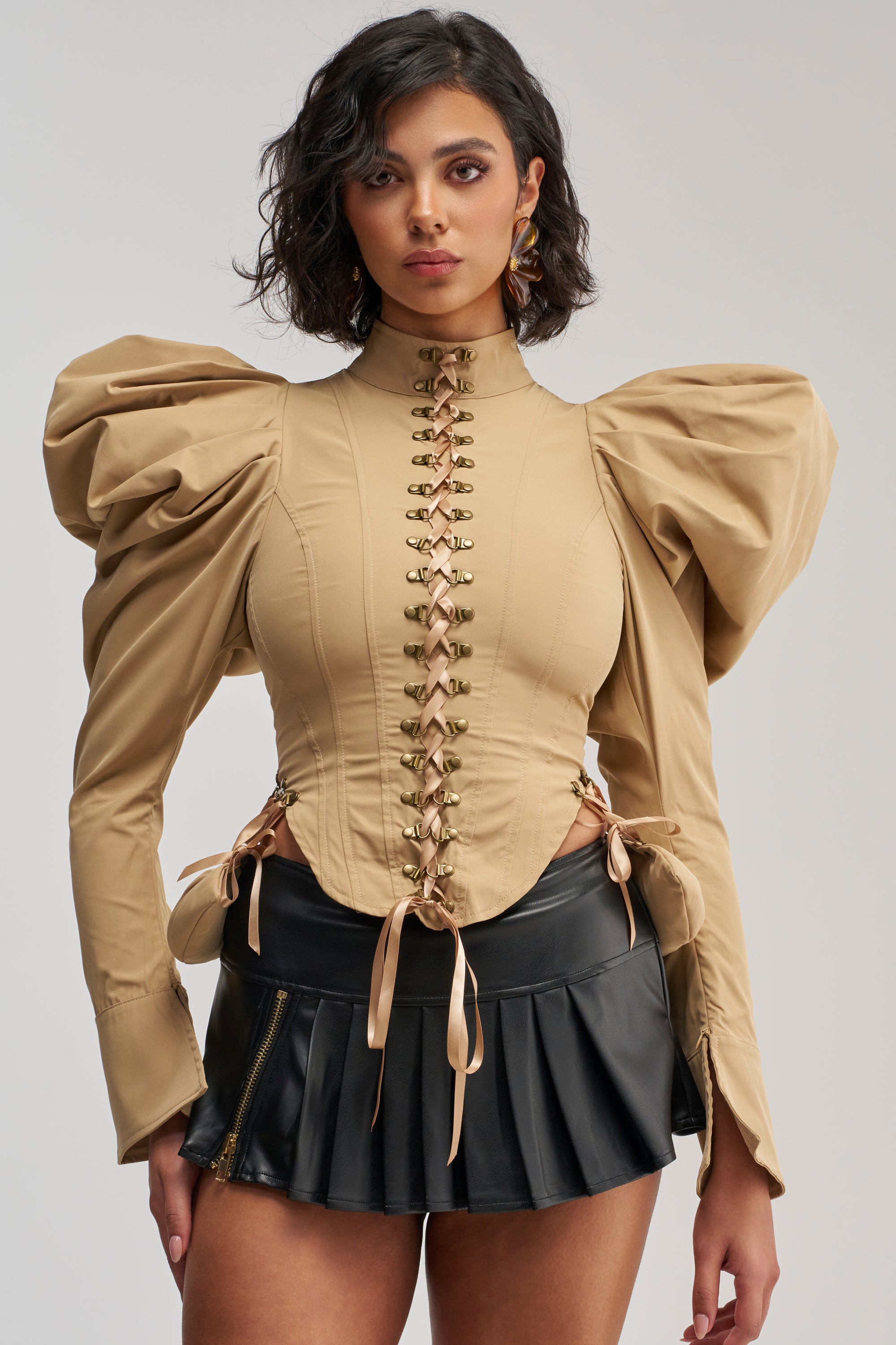 A woman wearing the NEW HEIGHTS POPLIN CORSET BLOUSE—featuring dramatic puffed sleeves, a lace-up front, and a khaki hue—paired with a black pleated mini skirt. She stands facing forward against a plain white background.