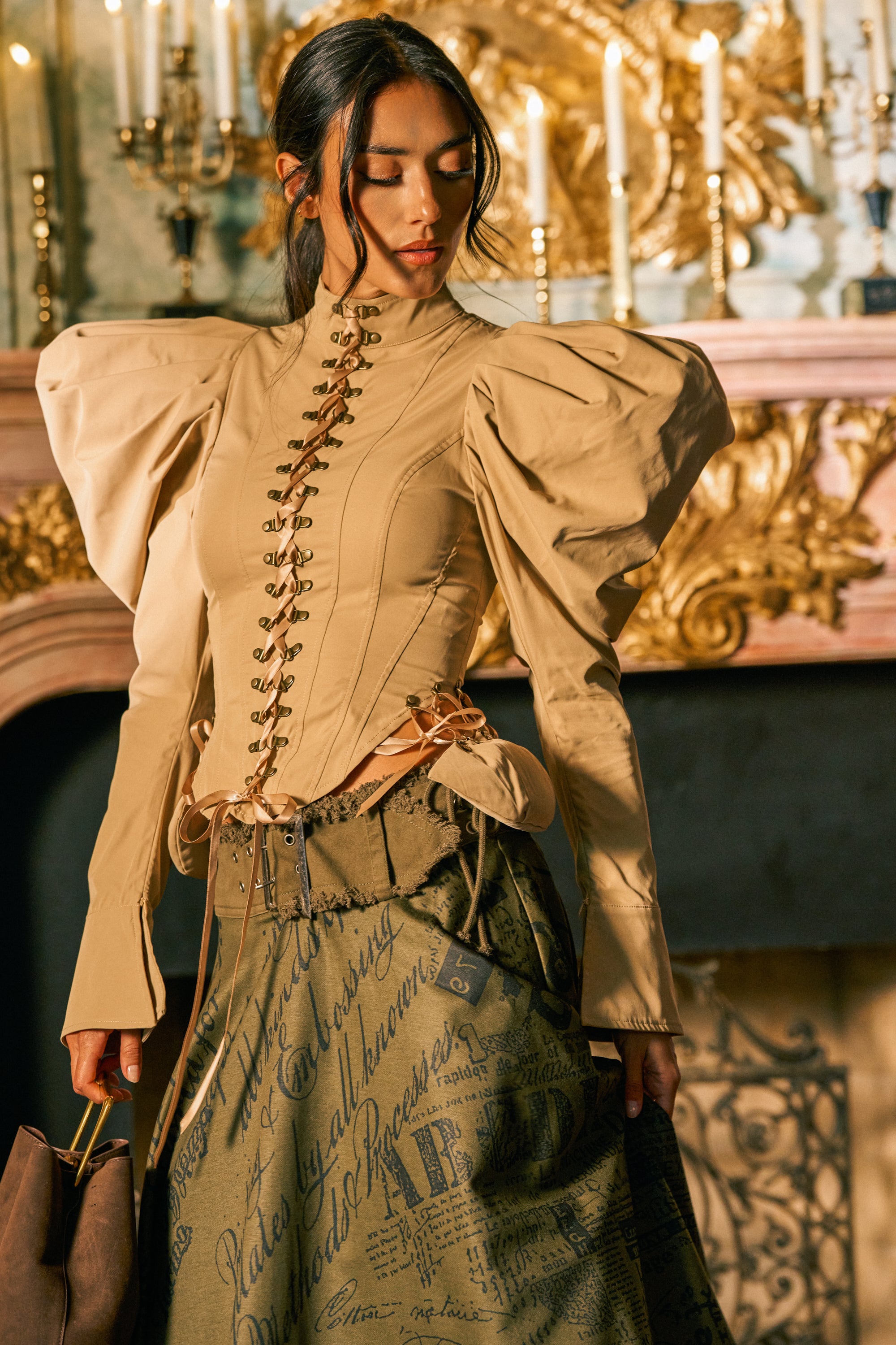 Wearing the NEW HEIGHTS POPLIN CORSET BLOUSE with vintage-inspired puff sleeves and a long printed skirt, a woman stands before a fireplace adorned with lit candles and ornate gold decor, showcasing statement fall styling.