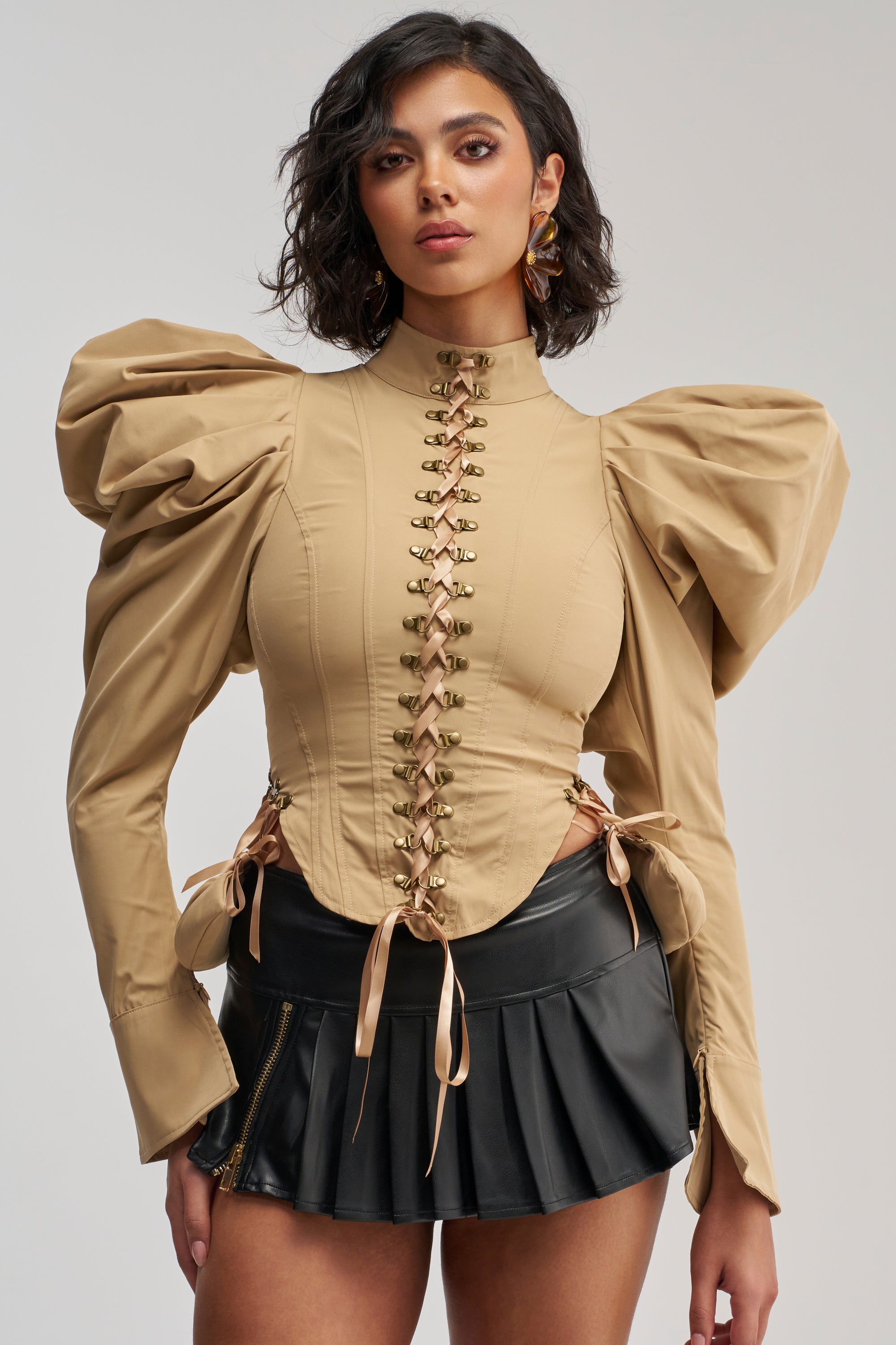 Wearing the NEW HEIGHTS POPLIN CORSET BLOUSE in khaki, featuring large puffed sleeves and a lace-up front, she poses against a plain light background, pairing the statement fall top with a short black pleated skirt accented by a zipper.