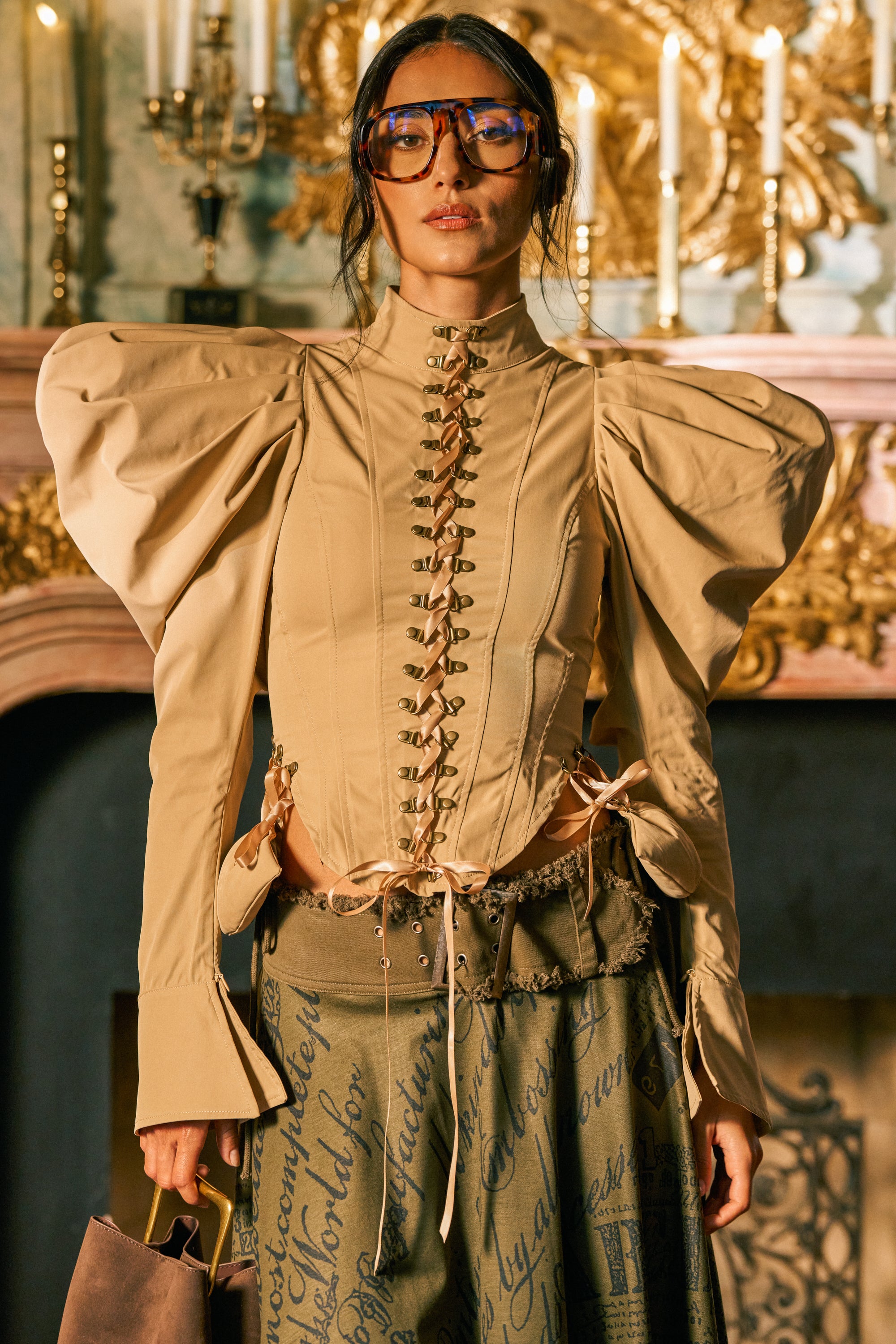 A woman stands indoors by an ornate fireplace with lit candles in the background, exemplifying statement fall styling perfect for chic date night looks, wearing blue glasses, a skirt with handwritten text, and the NEW HEIGHTS POPLIN CORSET BLOUSE featuring dramatic lace-up detailing and puff sleeves.