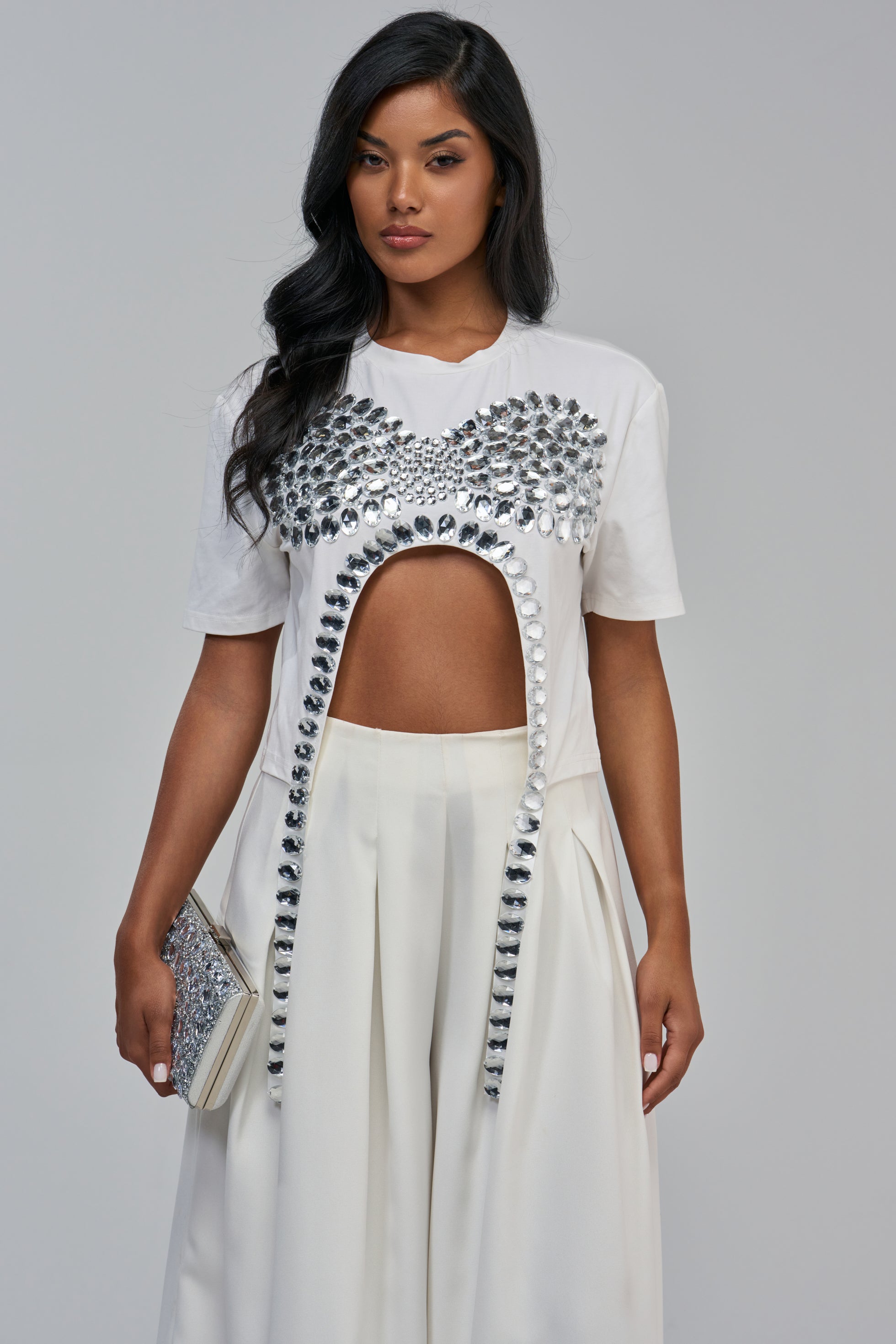 A woman wears the white IM THE GIFT RHINESTONE TEE featuring a large torso cutout framed by silver rhinestones, paired with white trousers and a matching rhinestone clutch.
