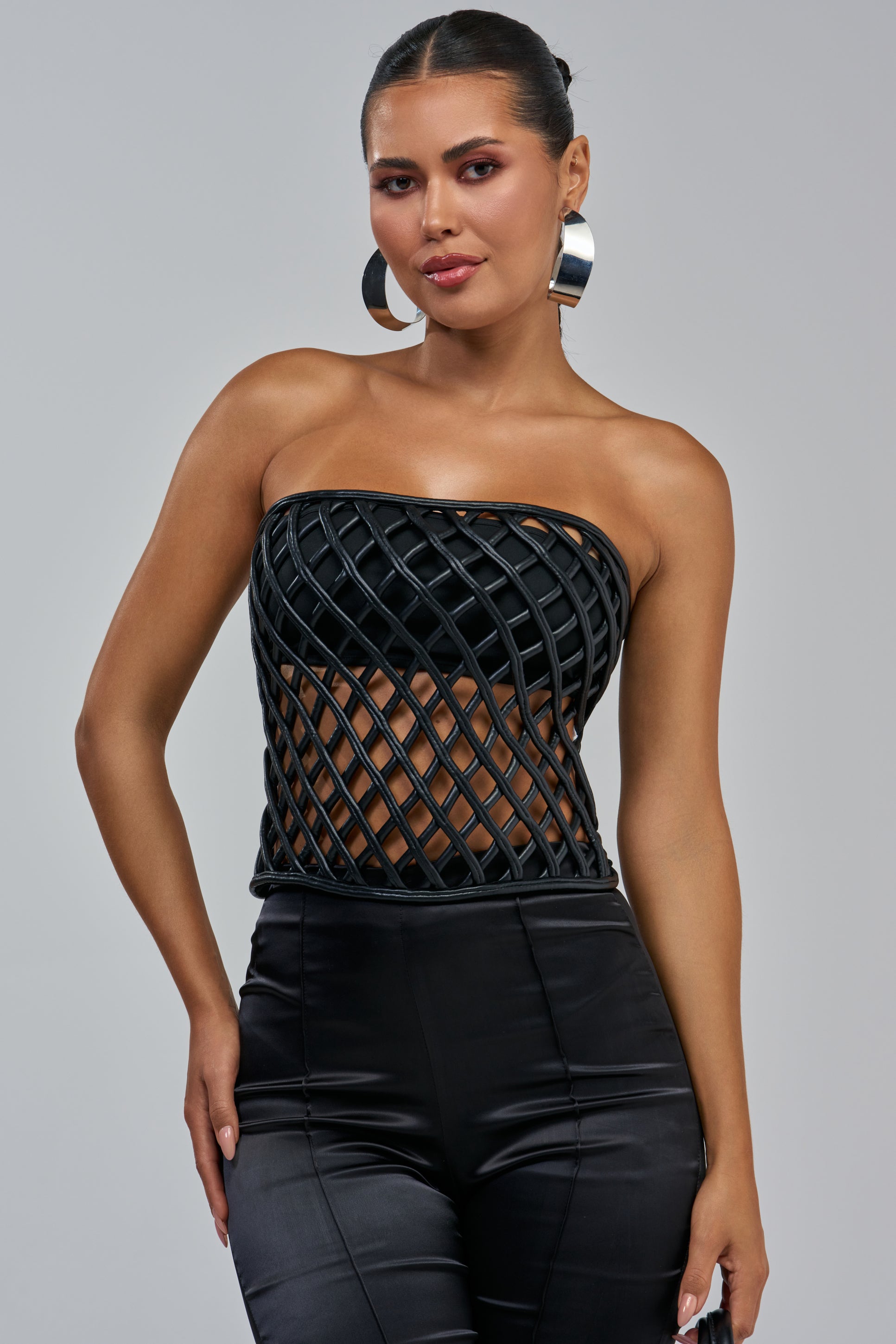 Woman wearing shiny black pants and the NAKIA FAUX LEATHER CORSET TOP with large hoop earrings, posing confidently against a plain gray background—perfect statement night out styling.