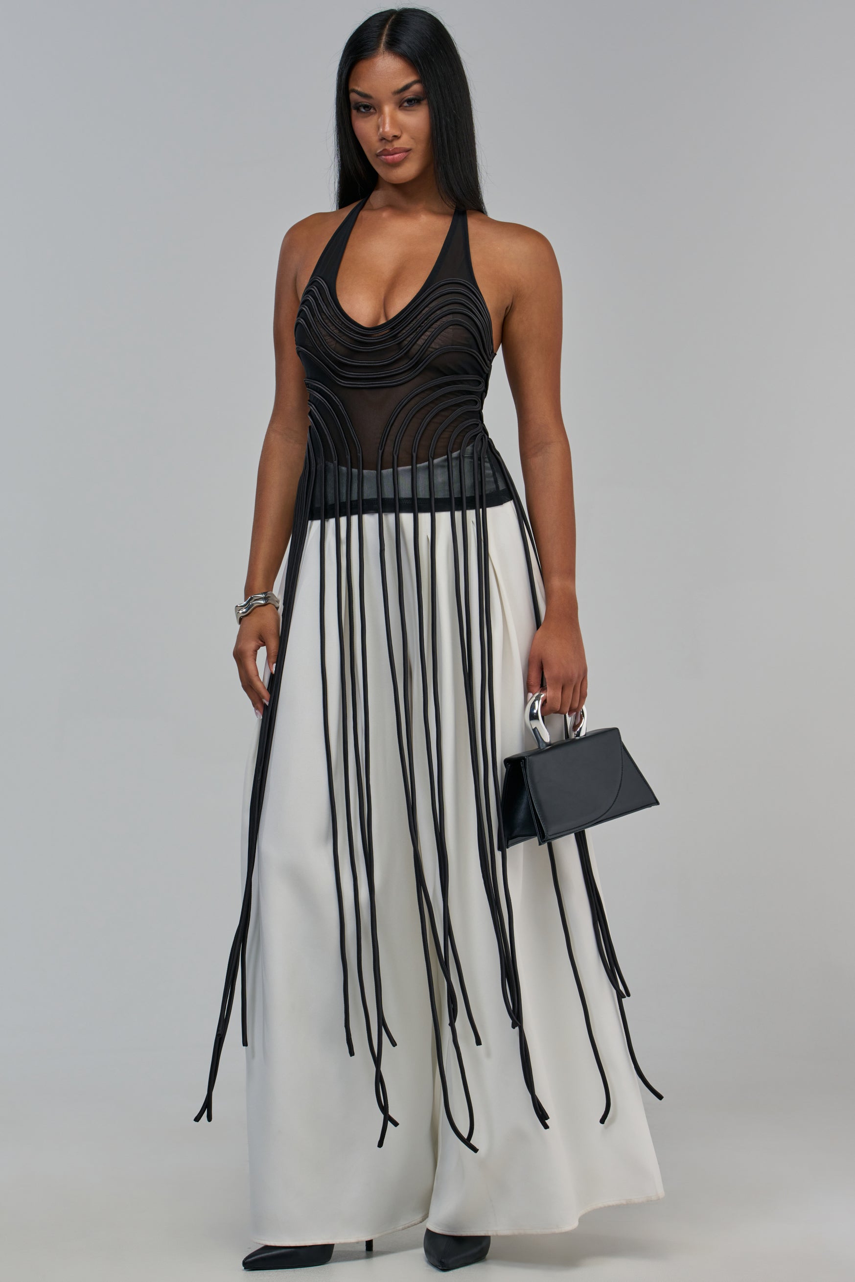 A woman wears a black DAYA MESH TOP with curved piping details and fringe, paired with wide-leg white trousers and holding a small black handbag.