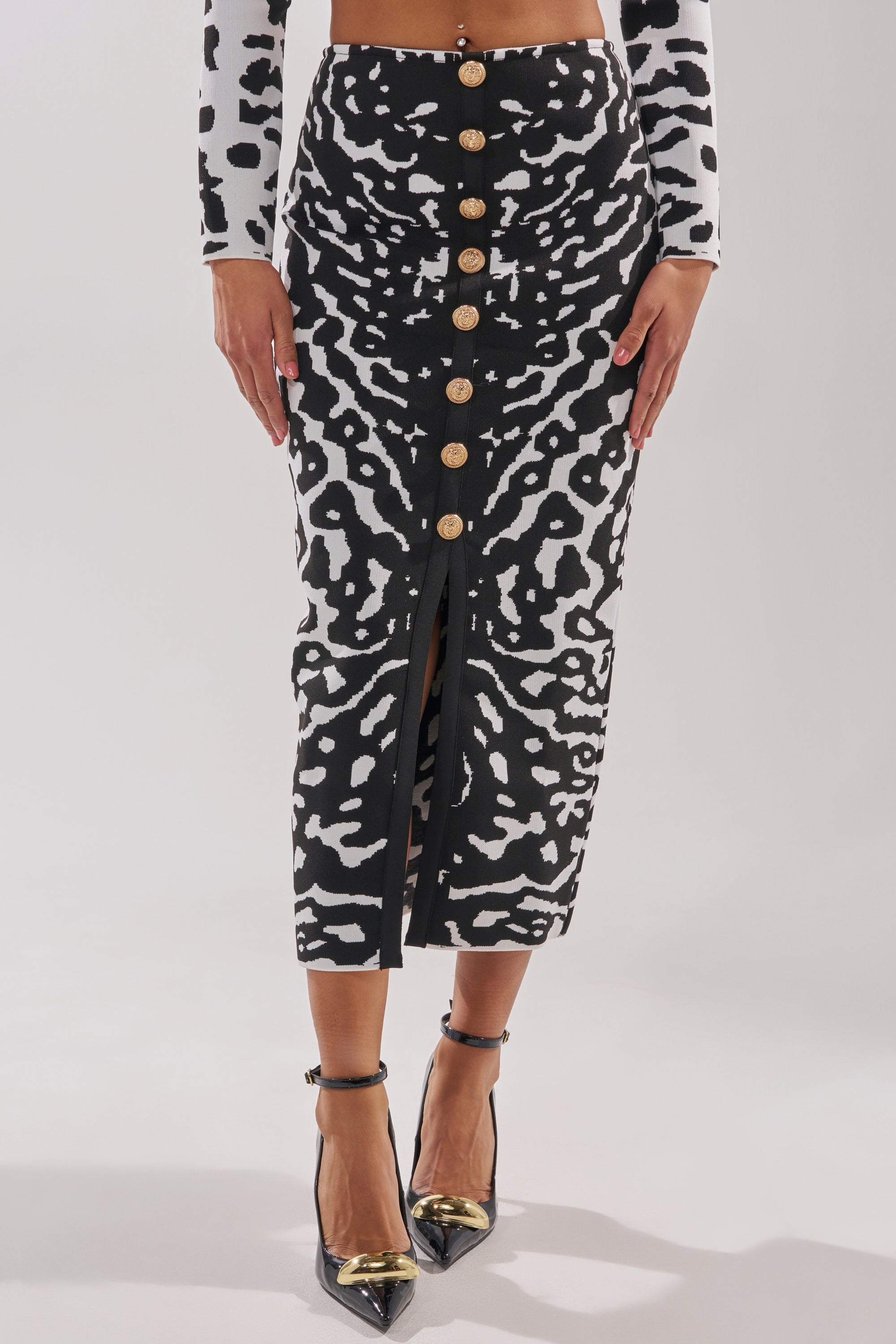 Wearing the BEST DRESSED MAXI SKIRT featuring a black and white abstract print, gold front buttons, and a front slit, paired with black pointed heels with gold tips and a coordinating long-sleeve top.