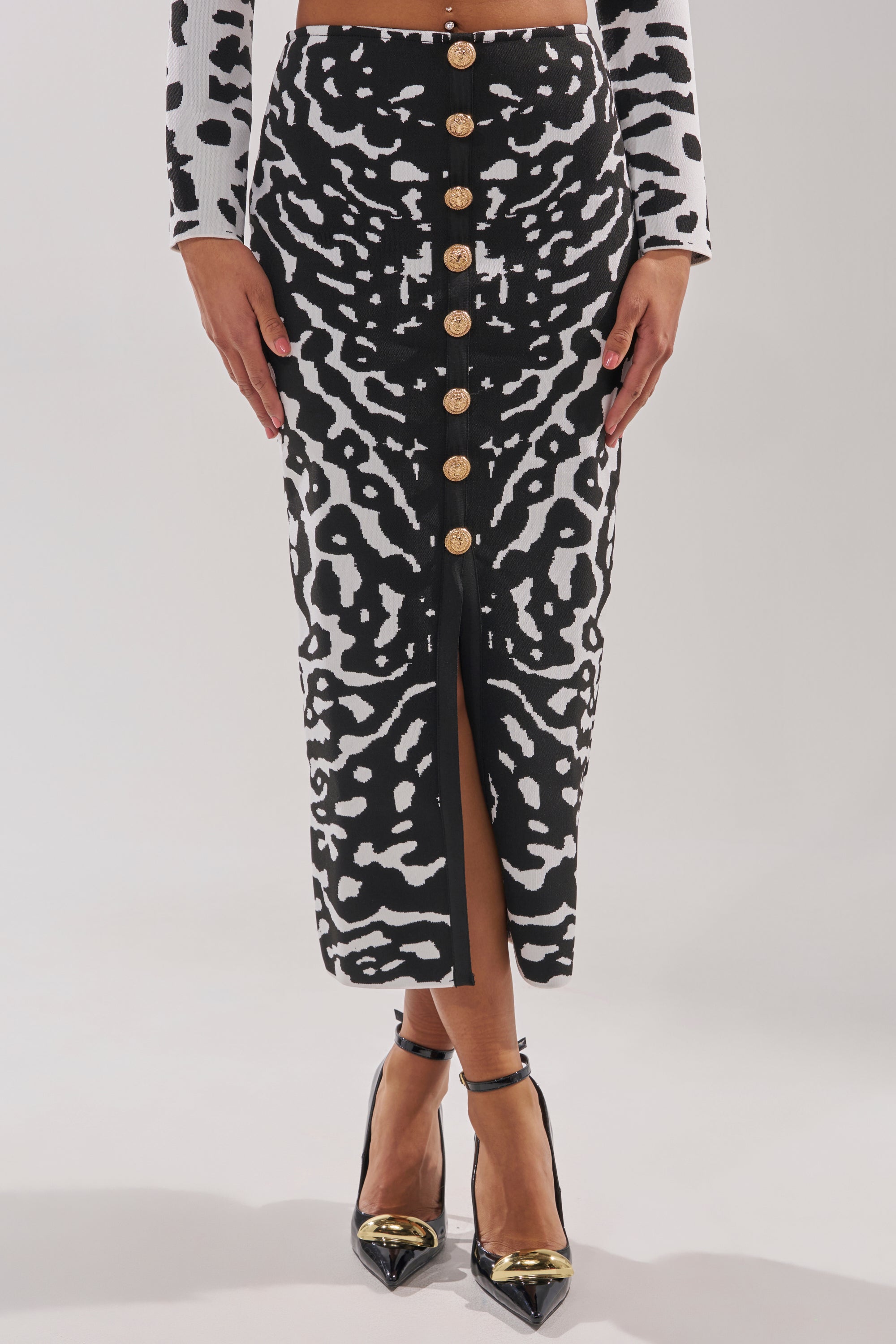A person wearing the BEST DRESSED MAXI SKIRT featuring a black and white abstract print with gold buttons, paired with a matching long-sleeve top and black pointed-toe heels with gold accents.