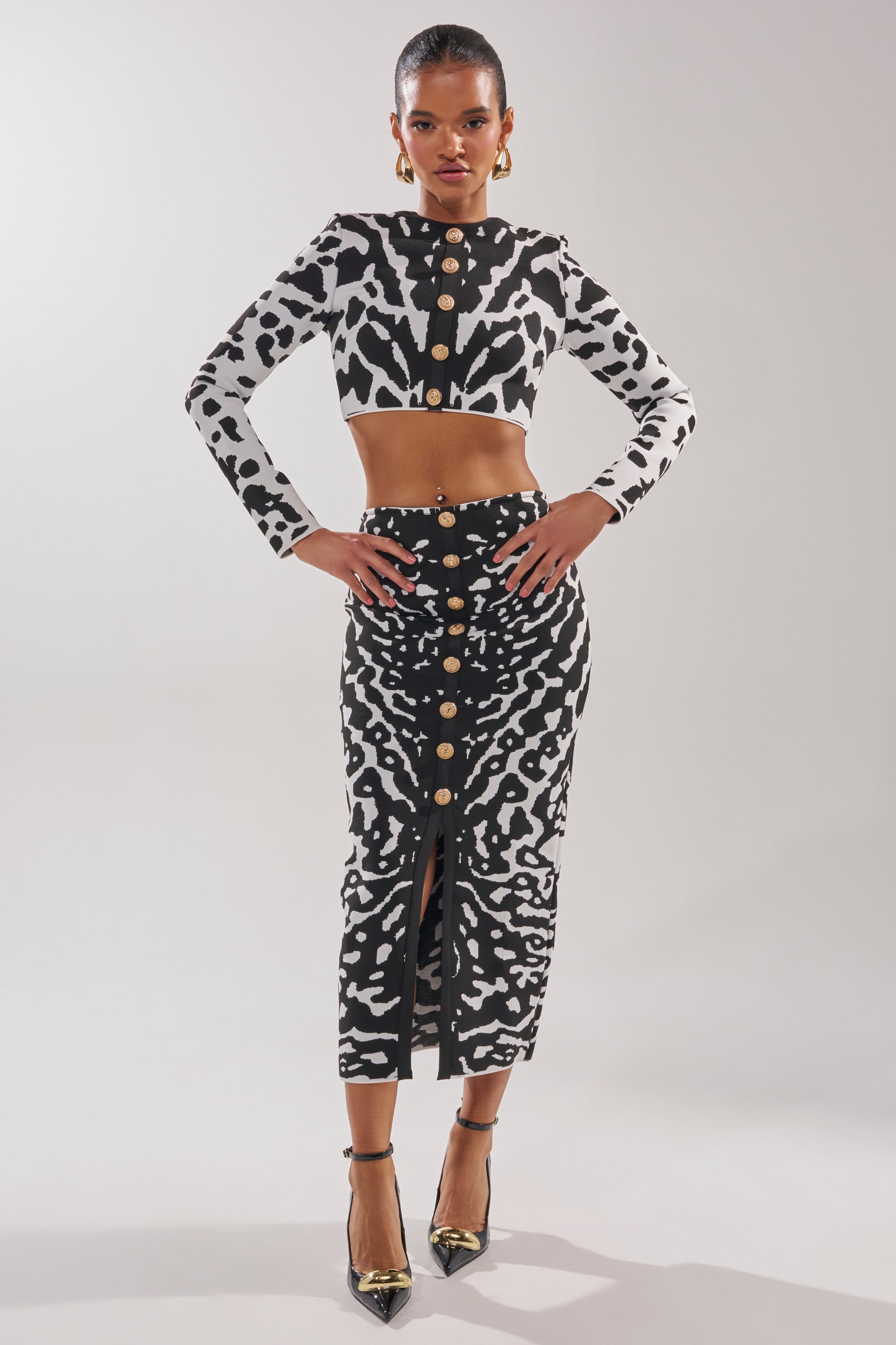 A woman stands confidently in the BEST DRESSED MAXI SKIRT, a high-rise black-and-white abstract print with gold front buttons. She pairs it with a matching crop top, slicked-back hair, gold earrings, and black pointed heels on a light background.