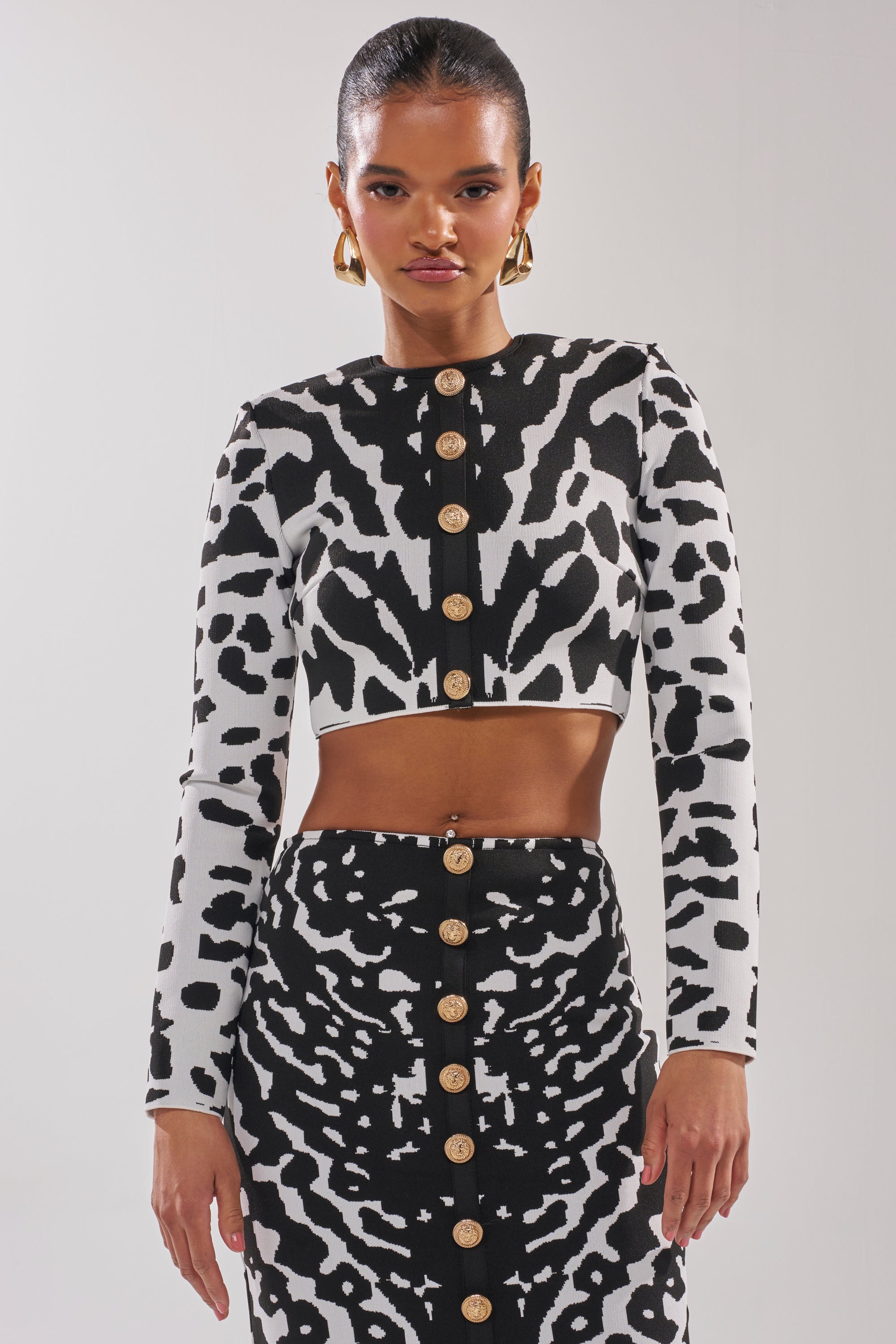 A woman exudes chic flair in a black-and-white animal print crop top and skirt, gold button accents, and hoop earrings—perfect inspiration for styling with the BEST DRESSED CARDIGAN’s abstract print. 0