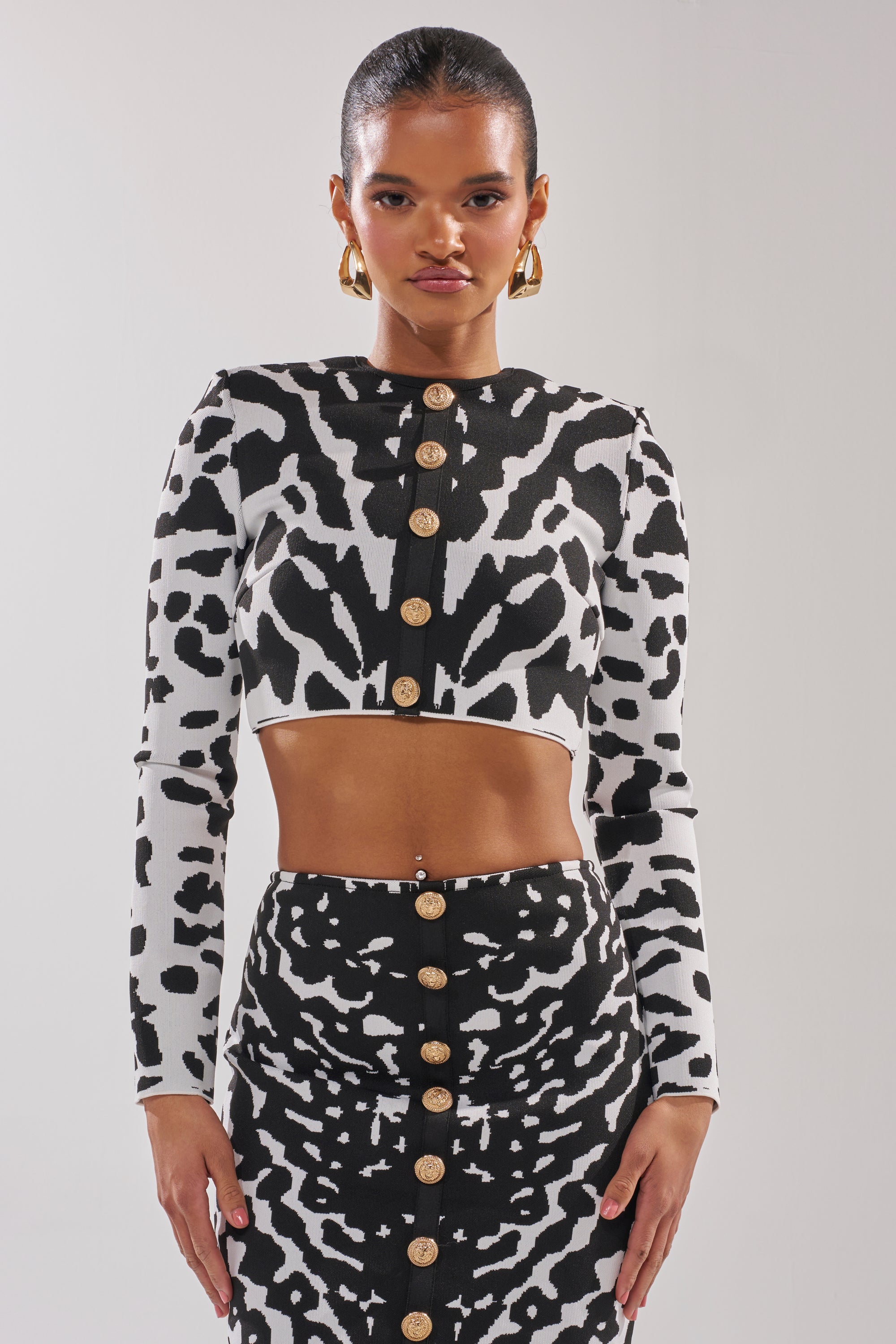 A woman wears a black and white animal print crop top and skirt with gold buttons, layering with the BEST DRESSED CARDIGAN. She accessorizes with gold hoop earrings and styles her hair slicked back. 1