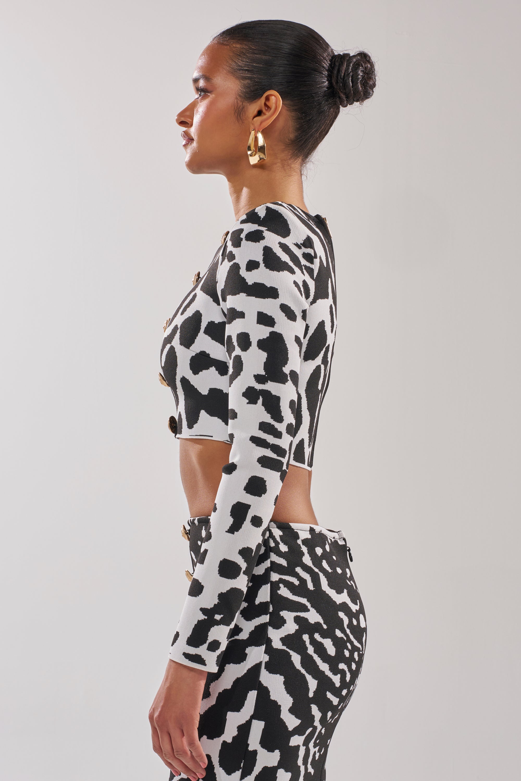 A woman with her hair in a bun and gold hoop earrings models the BEST DRESSED CARDIGAN over a matching black-and-white animal print crop top and skirt, standing in profile against a plain light background. 2