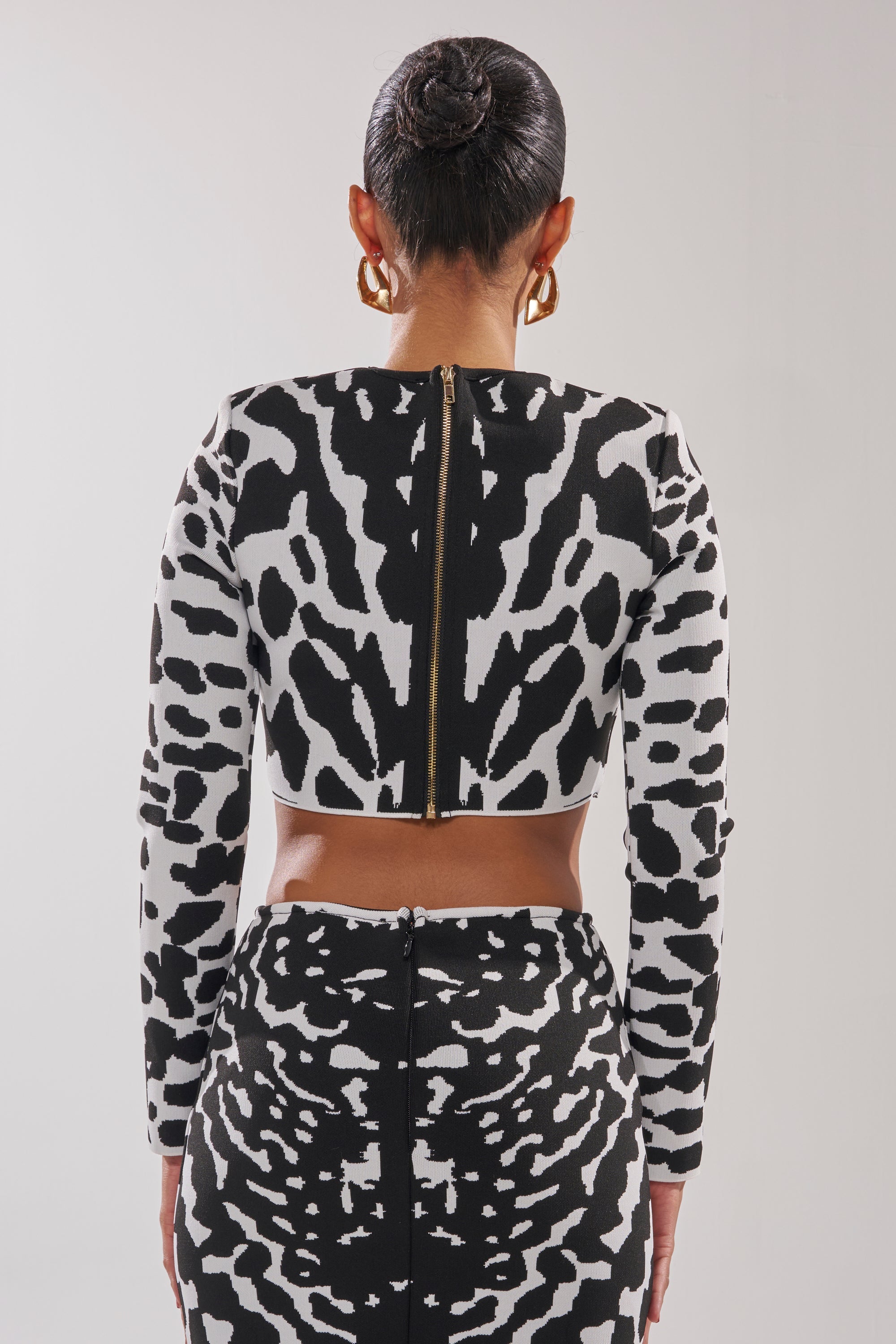 A woman with her hair in a sleek bun and large gold hoop earrings faces away from the camera, wearing the BEST DRESSED CARDIGAN over a black and white animal print skirt. 3