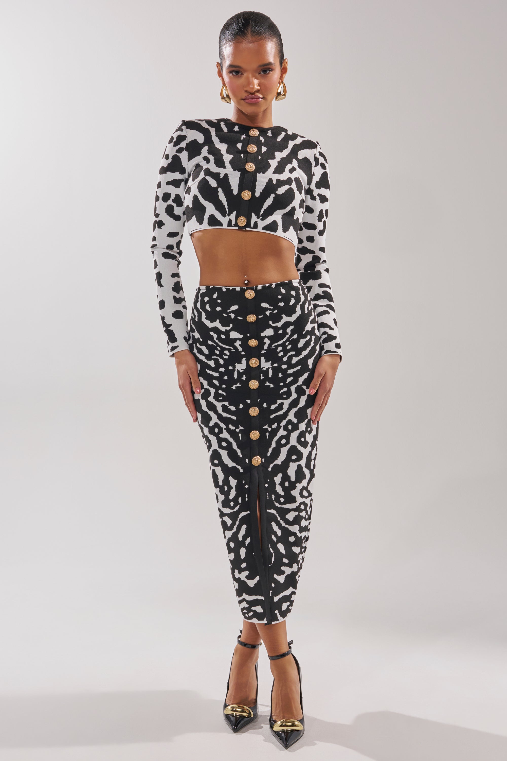 A woman poses against a plain background in the BEST DRESSED CARDIGAN, styled with a black-and-white animal print crop top, coordinated high-waisted skirt featuring gold buttons, black pointed heels, and gold earrings. 4