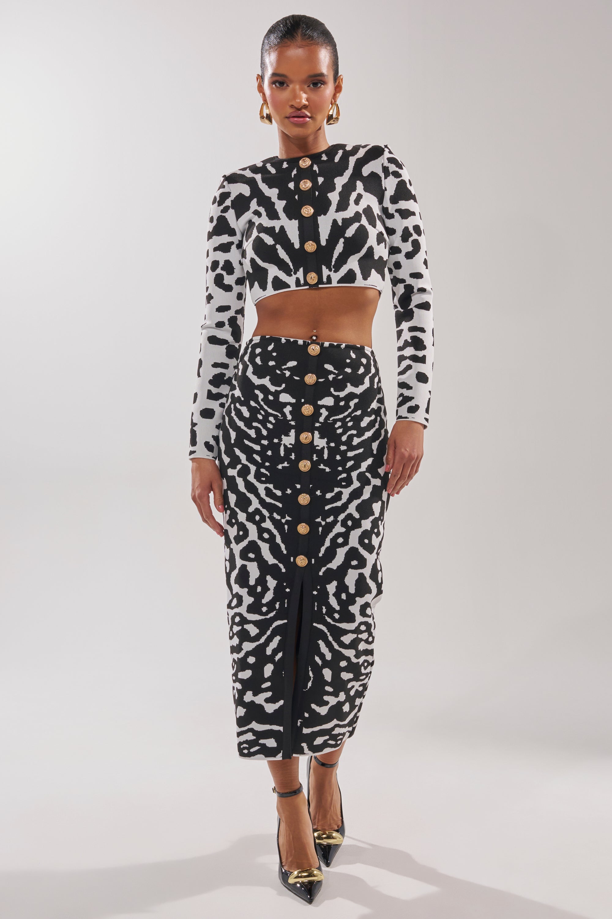 A woman faces forward in a black and white animal print crop top with gold buttons and a high-waisted ankle-length skirt. She completes the look with black heels, gold earrings, and the BEST DRESSED CARDIGAN draped over her shoulders. 6