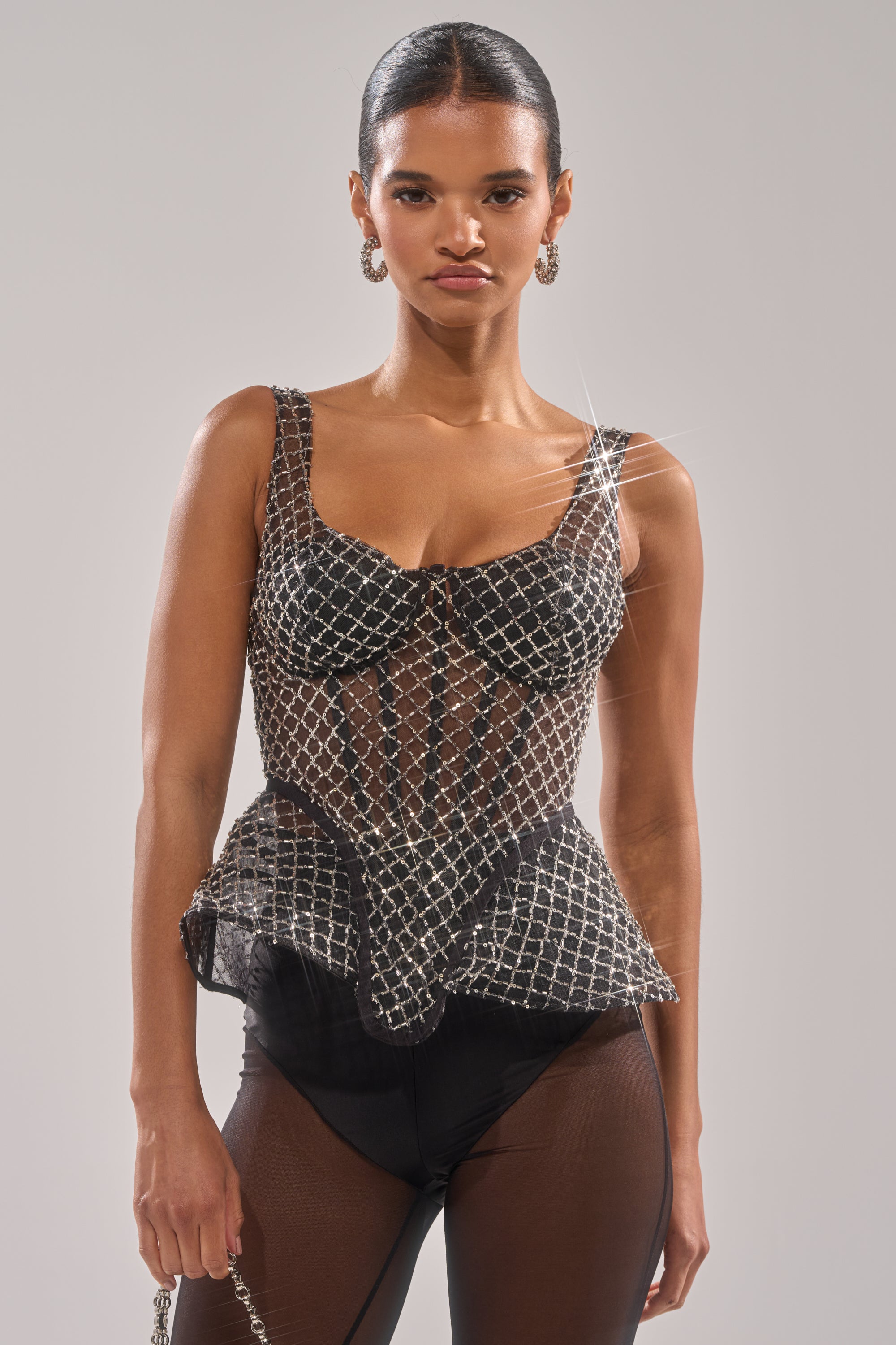 A woman with slicked-back hair wears the GLITZ AND GLAM SHEER RHINESTONE CORSET—a sleeveless, fitted, semi-sheer black top with a sparkling diamond-pattern overlay—paired with dark high-waisted bottoms against a plain light background. 1
