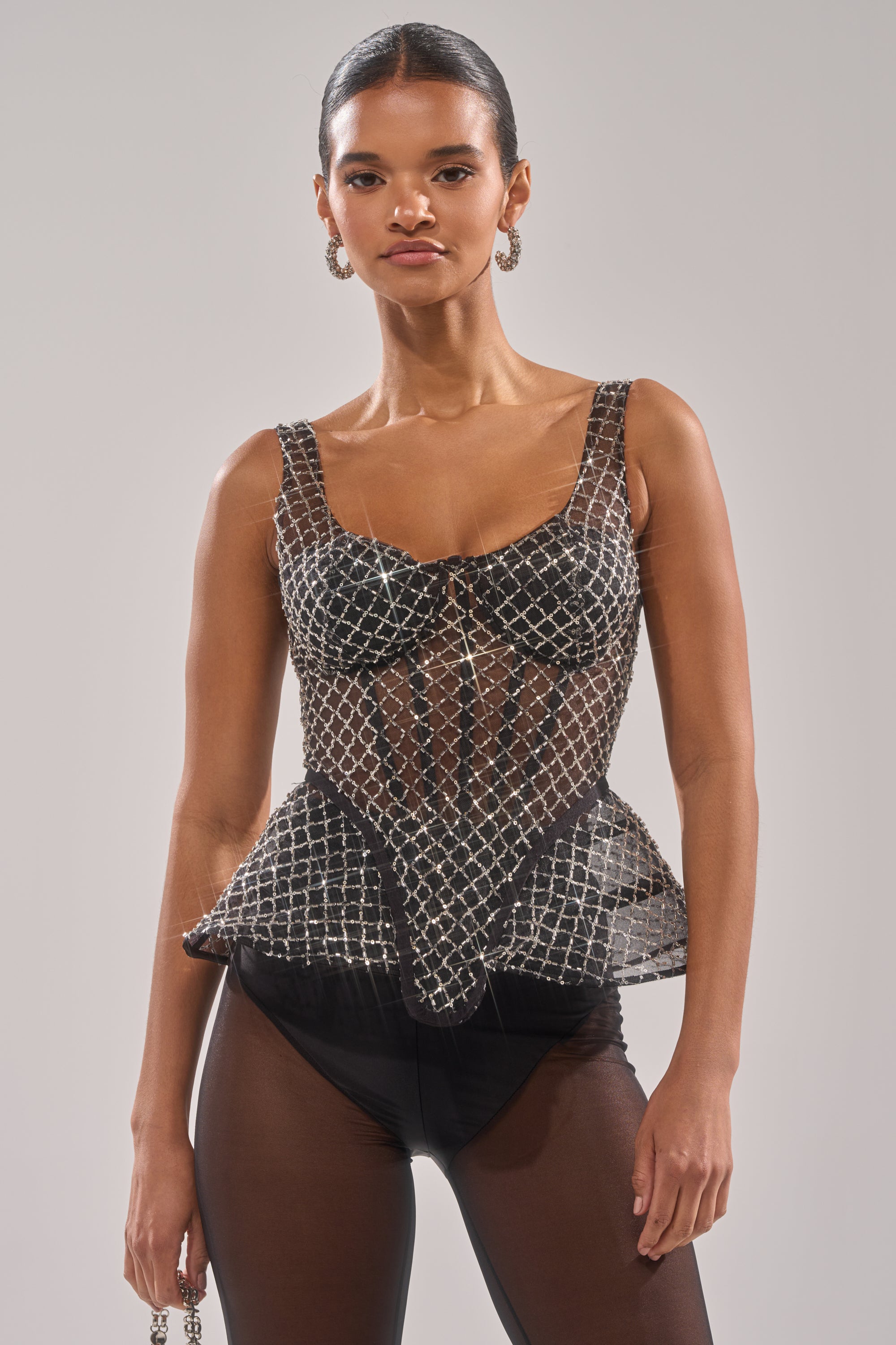 A person models the GLITZ AND GLAM SHEER RHINESTONE CORSET with a crisscross silver pattern, paired with black bottoms and hoop earrings, standing against a plain light background. 2