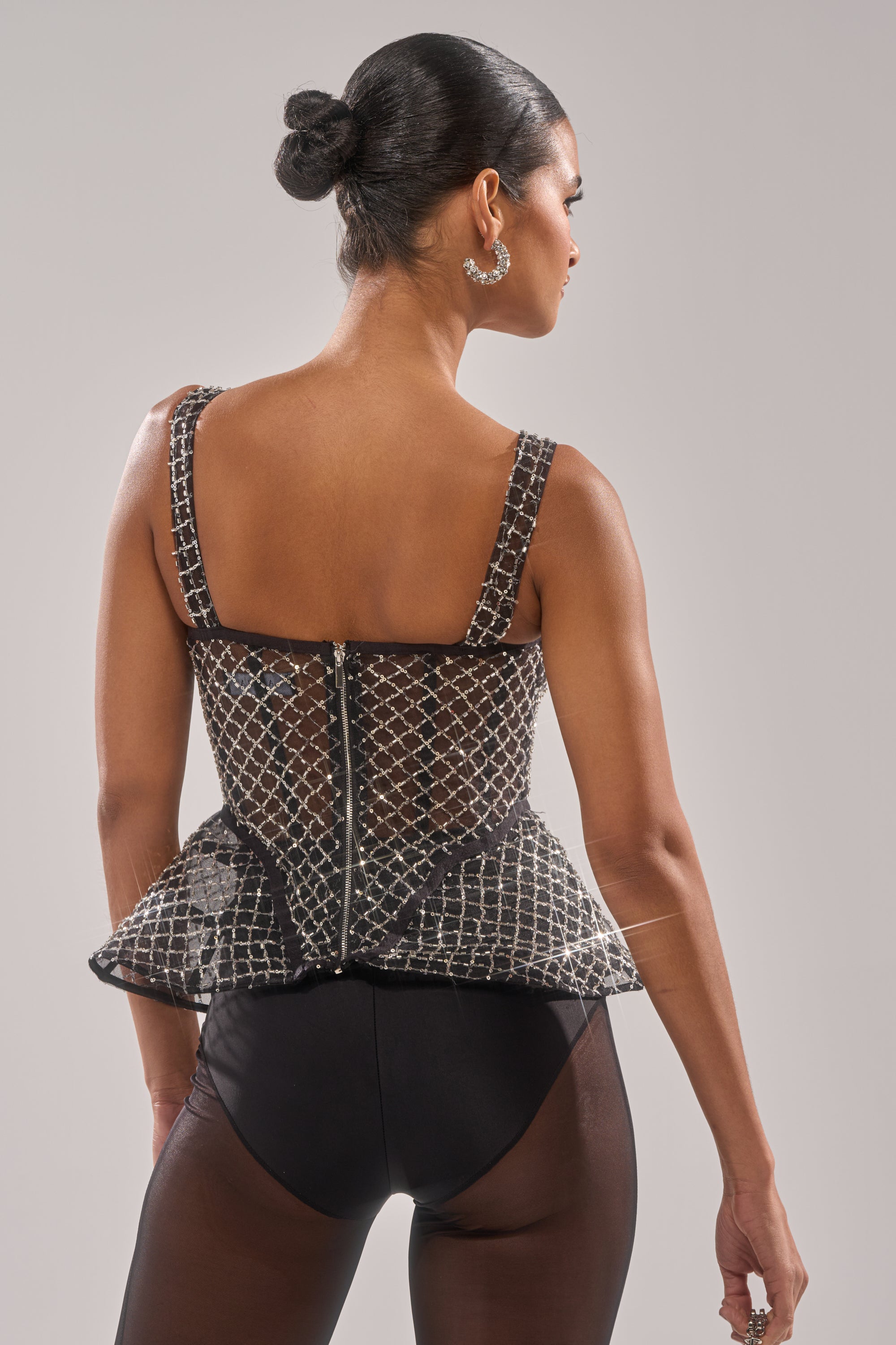 A woman with dark hair in a sleek bun models the GLITZ AND GLAM SHEER RHINESTONE CORSET, paired with black bottoms. Facing away, she showcases the back zipper detail and hoop earrings for a rocker chic vibe against a plain backdrop. 5