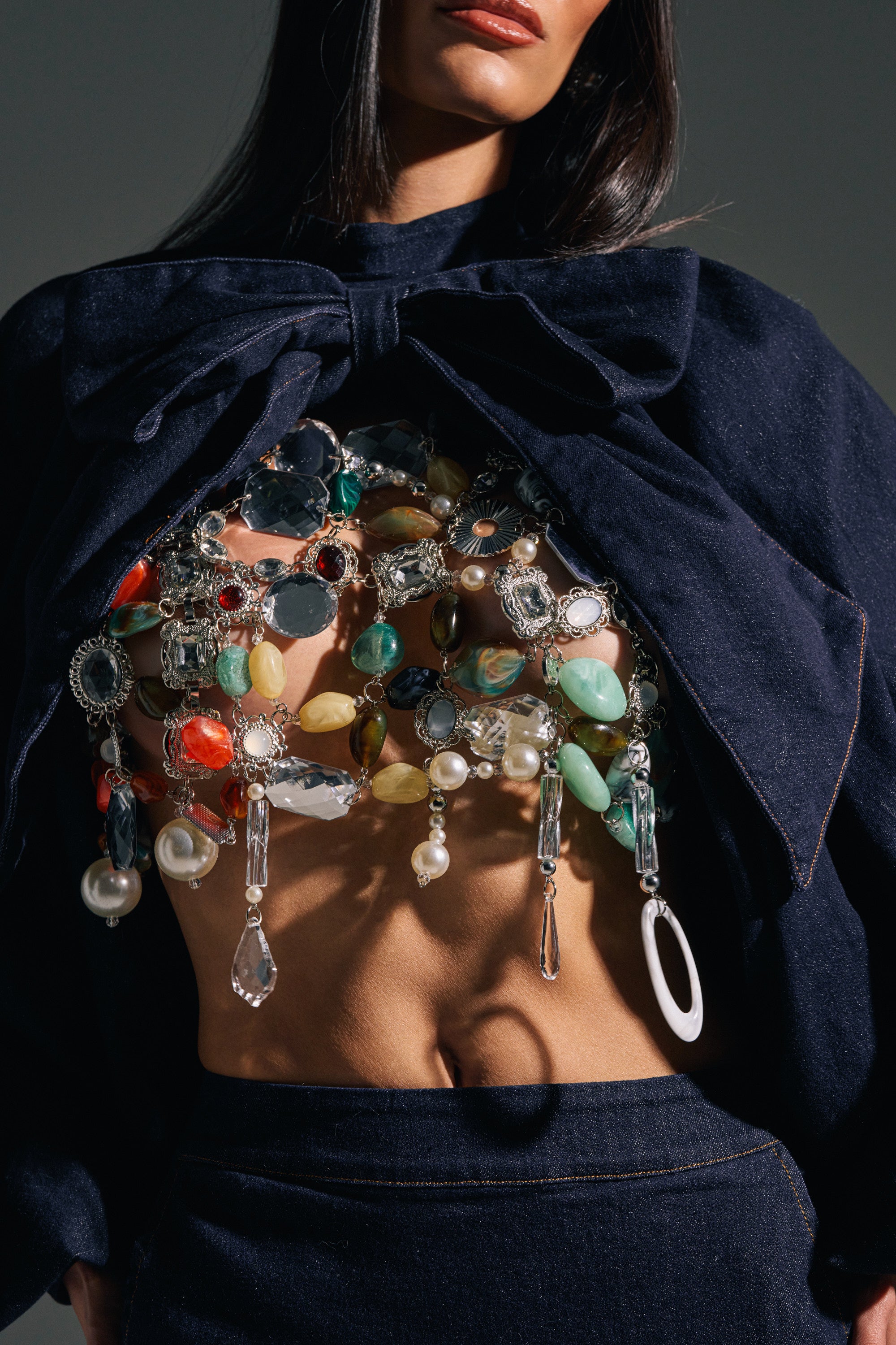 A woman wears the SHAKE IT QUEEN BEADED BRALETTE, a dark denim halter with a large bow, revealing her midriff and adorned with colorful chunky gemstone and crystal details—a bold statement piece.
