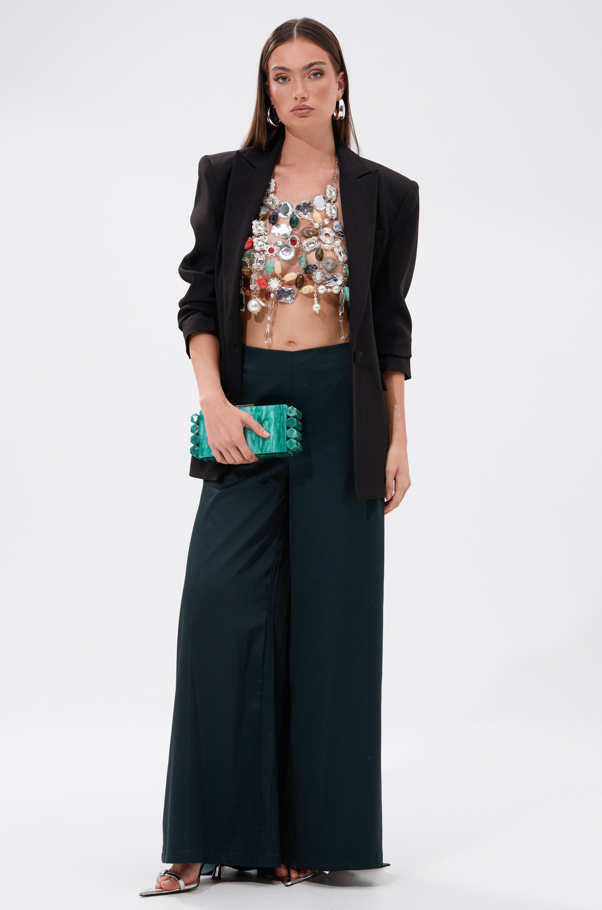 A woman wears the SHAKE IT QUEEN BEADED BRALETTE under a black blazer with wide-leg dark trousers and heeled sandals. She carries a green and black clutch, her look polished and chic against a plain white background.