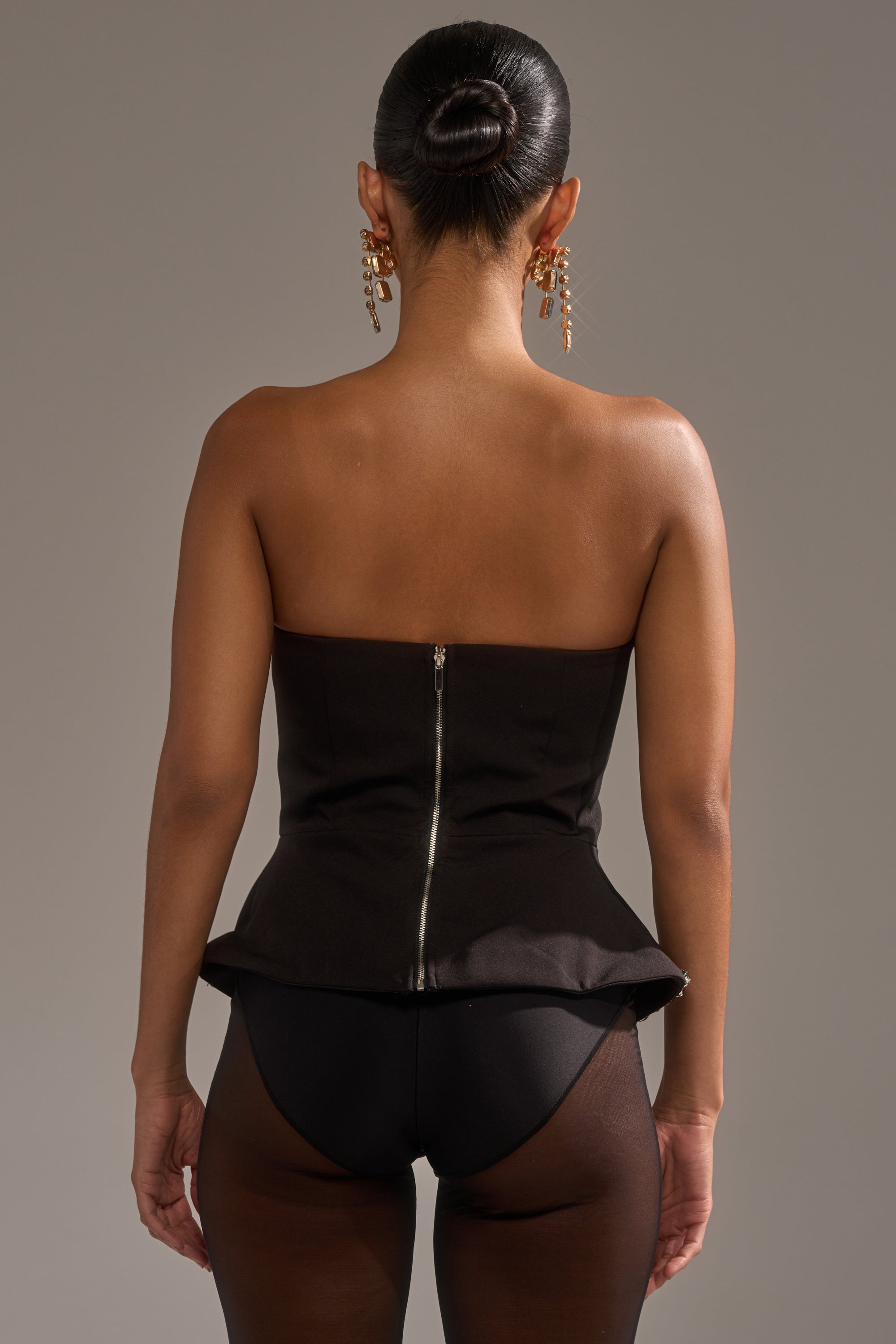 A woman, her hair in a sleek bun, faces away wearing the Jewels For Everyone Corset with a visible back zipper, sheer black tights, and gold dangling earrings against a neutral backdrop.