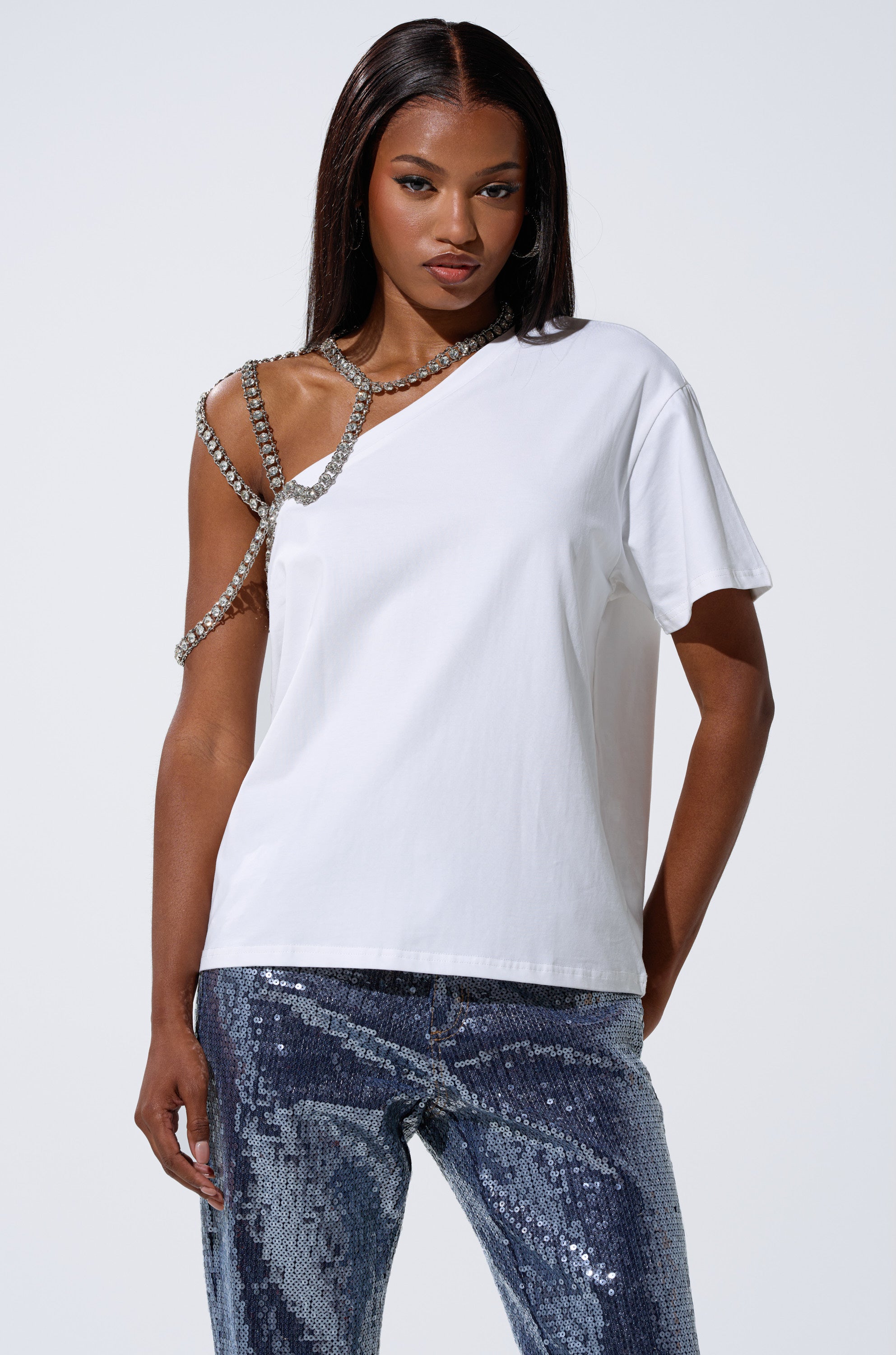 A woman wears the DANCING STAR OVERSIZED TEE, featuring an asymmetrical fit and beaded chain detail on one shoulder, styled with blue sequined pants and a rhinestone harness, against a plain light background. 0