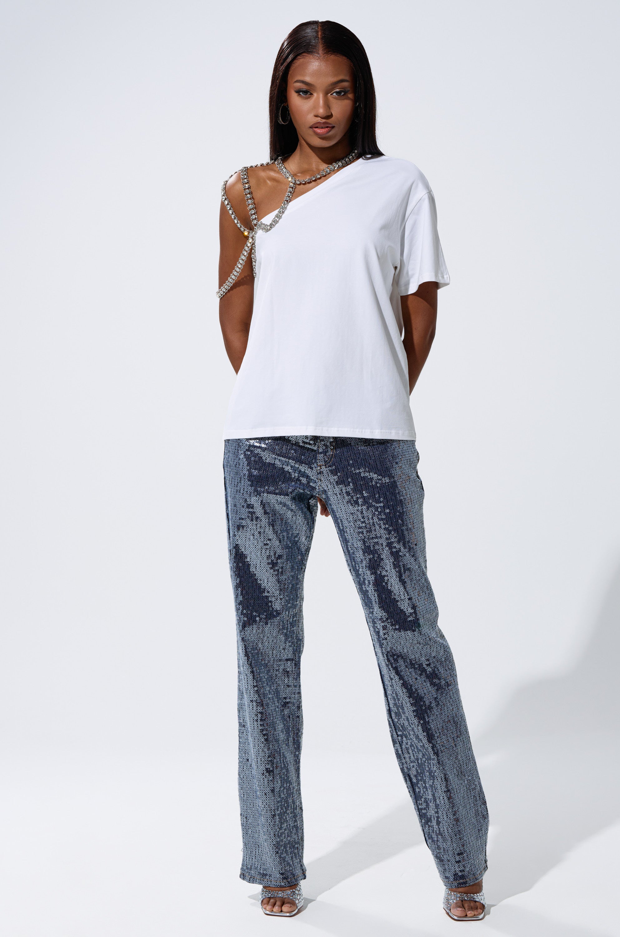 A woman faces forward in the DANCING STAR OVERSIZED TEE styled with a rhinestone harness, silver sequin pants, hoop earrings, open-toed heels, and long straight hair, set against a plain white background. 6