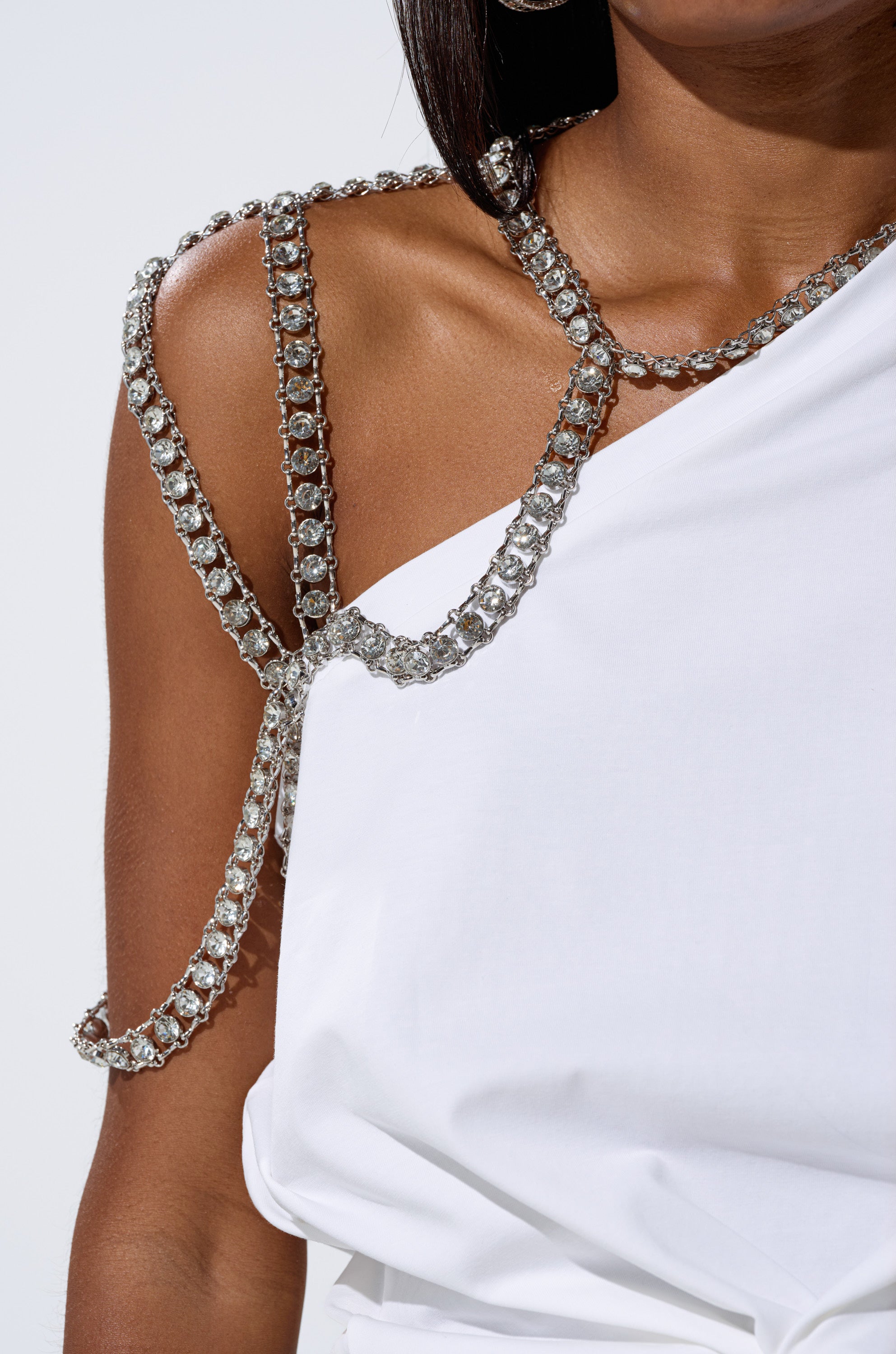 A person wears the white DANCING STAR OVERSIZED TEE with an asymmetrical fit and one-shoulder design, styled with sparkling silver chain jewelry and a rhinestone harness. The background is plain and minimalistic. 7