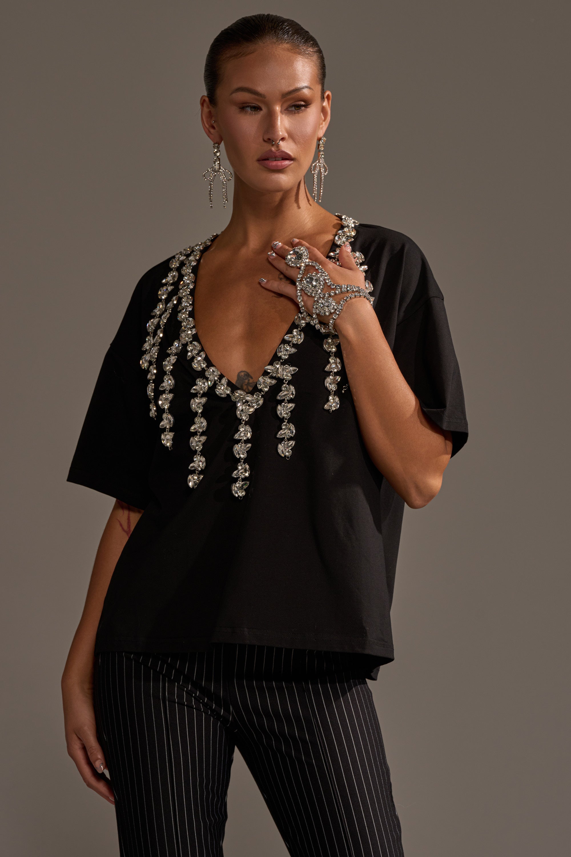 A woman wears the black RHINESTONE QUEEN OVERSIZED TEE adorned with sparkling silver rhinestones, paired with black pinstripe pants and ornate silver jewelry, standing against a neutral background.