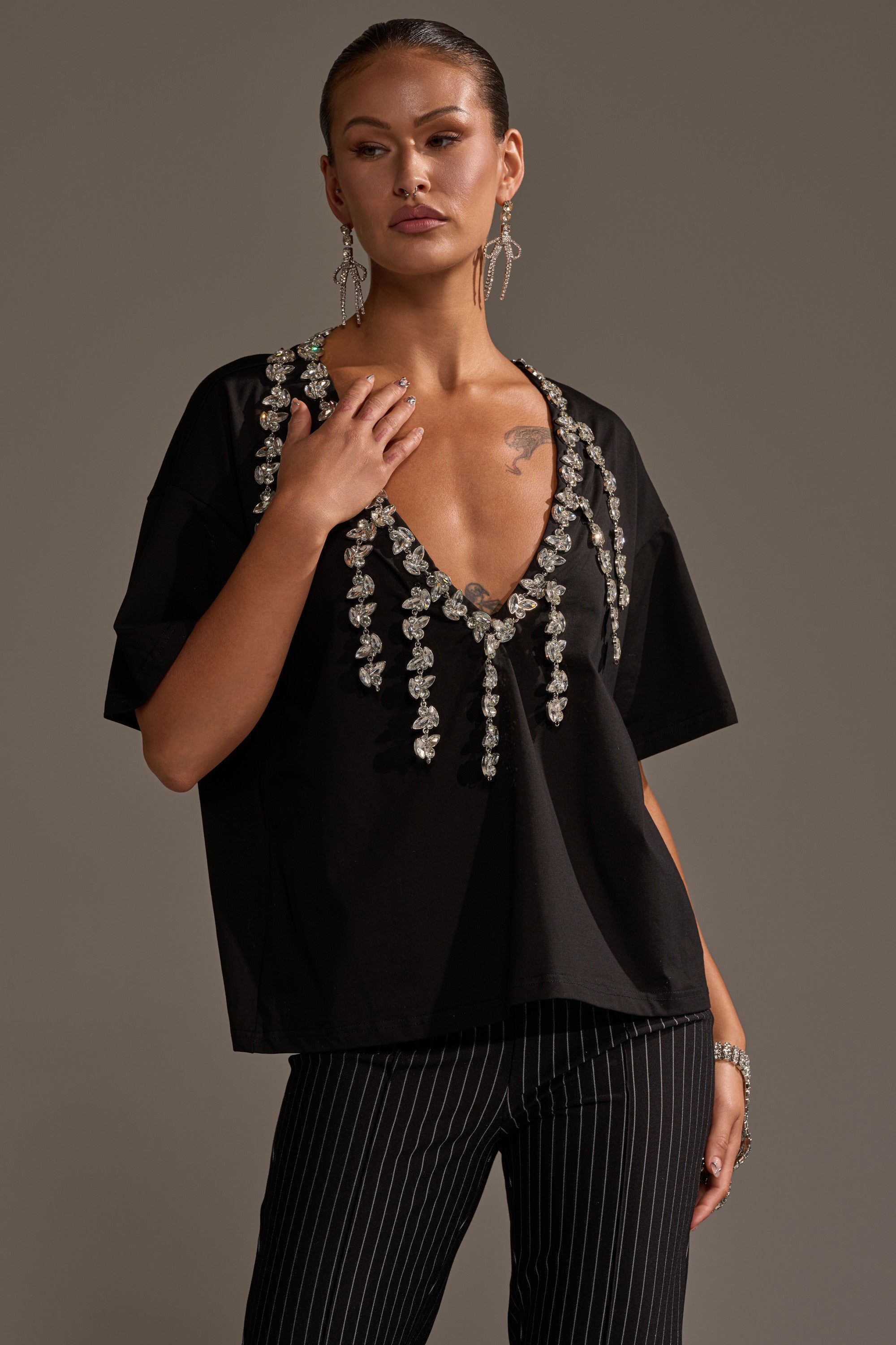 A woman poses against a plain brown background wearing the RHINESTONE QUEEN OVERSIZED TEE with a plunging V-neckline, pinstripe black pants, statement earrings, and a bracelet.