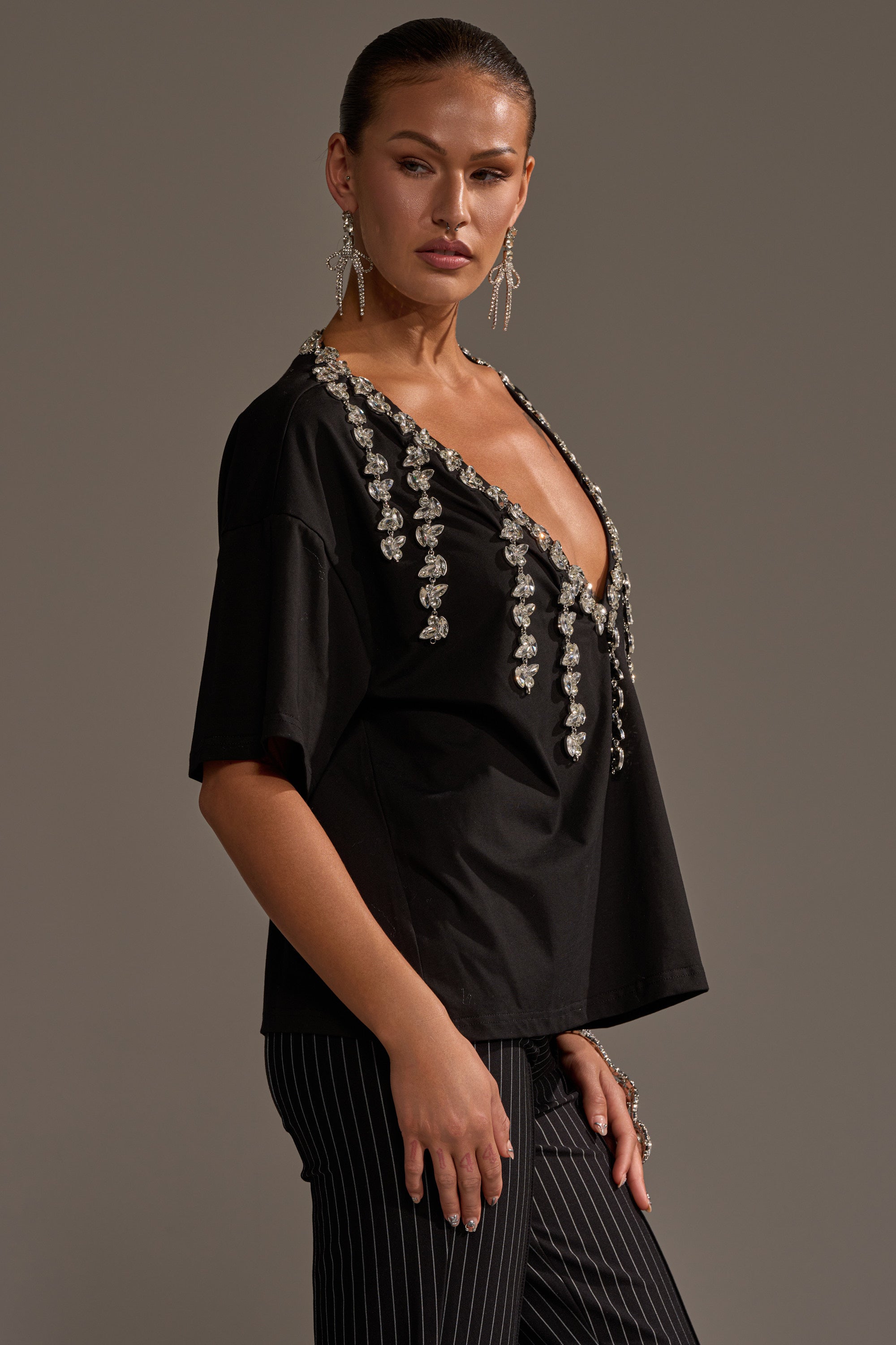 A woman in profile wears the RHINESTONE QUEEN OVERSIZED TEE with a plunging V-neck and black pinstripe pants. Her slicked-back hair and bold earrings highlight her neutral look against a plain gray backdrop.