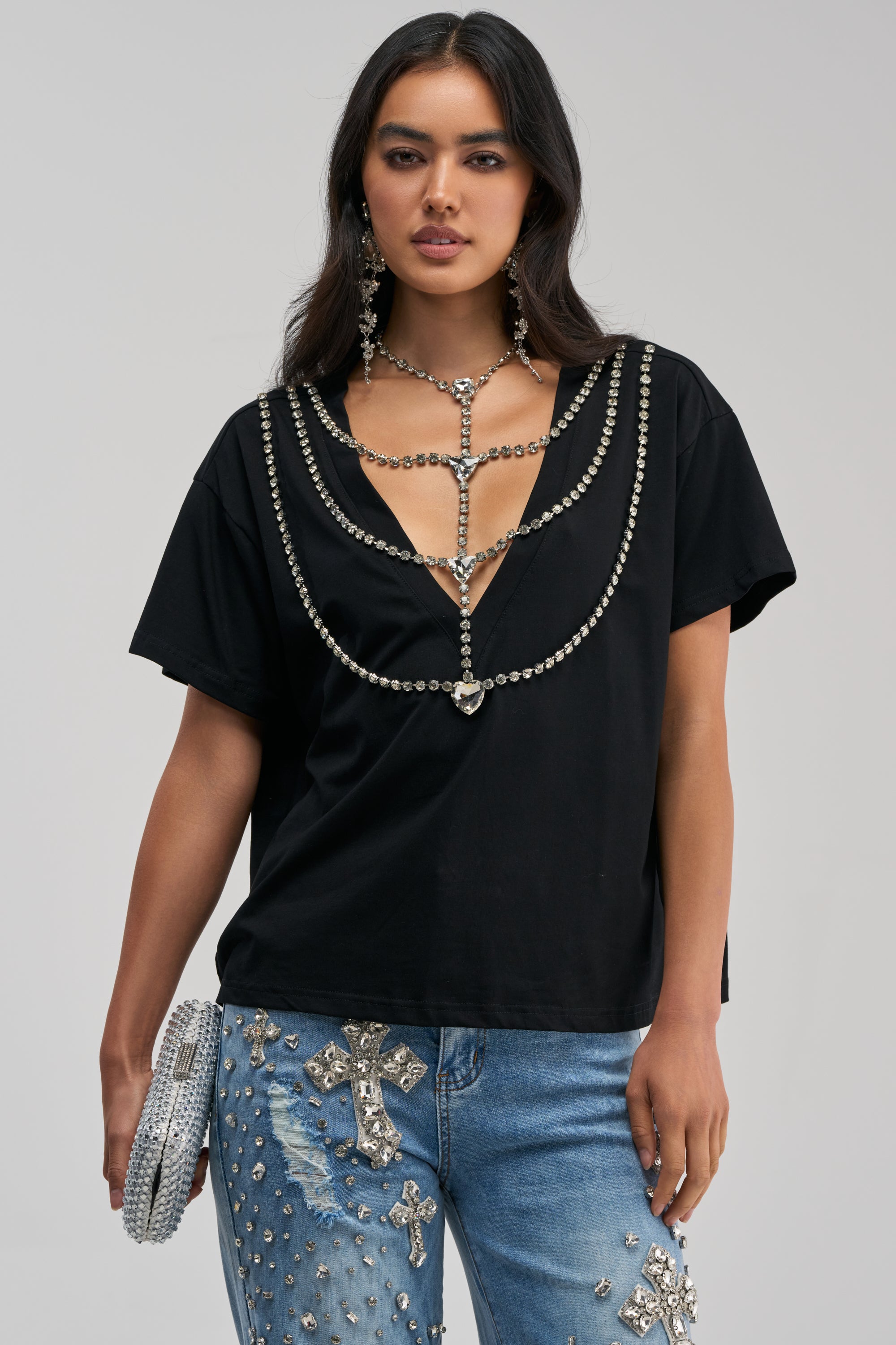 A woman models the DIAMONDS DANCING RHINESTONE DETAIL TEE IN BLACK, styled with blue rhinestone-embellished jeans and cross motifs, a silver clutch, and statement jeweled earrings—an eye-catching streetwear look ideal for concert-ready flair.