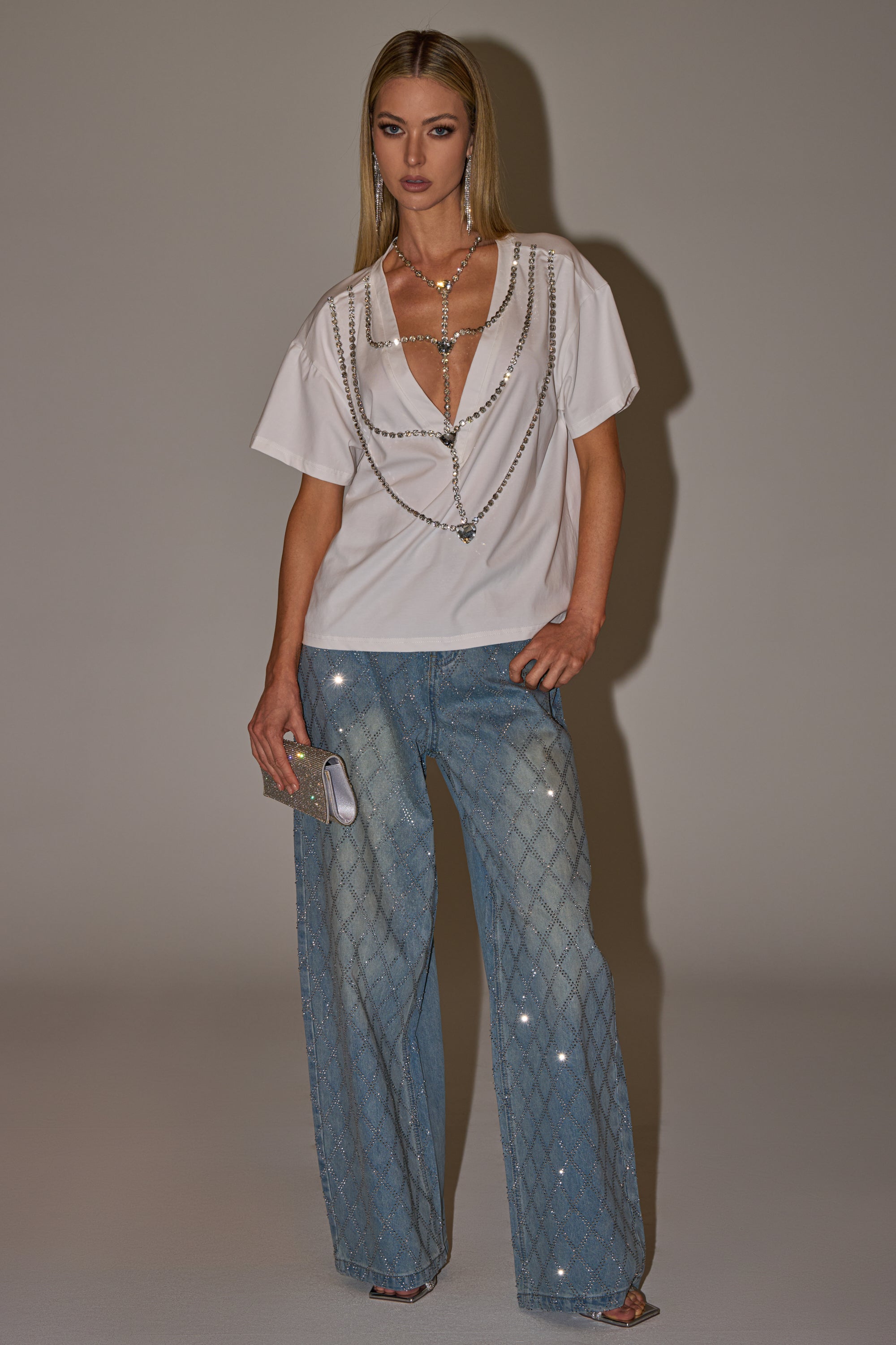 A woman wears the DIAMONDS DANCING RHINESTONE DETAIL TEE, featuring sparkling chain-draped sleeves, with high-waisted light blue diamond-pattern jeans, a glittery clutch, strappy heels, and layered jewelry. 9