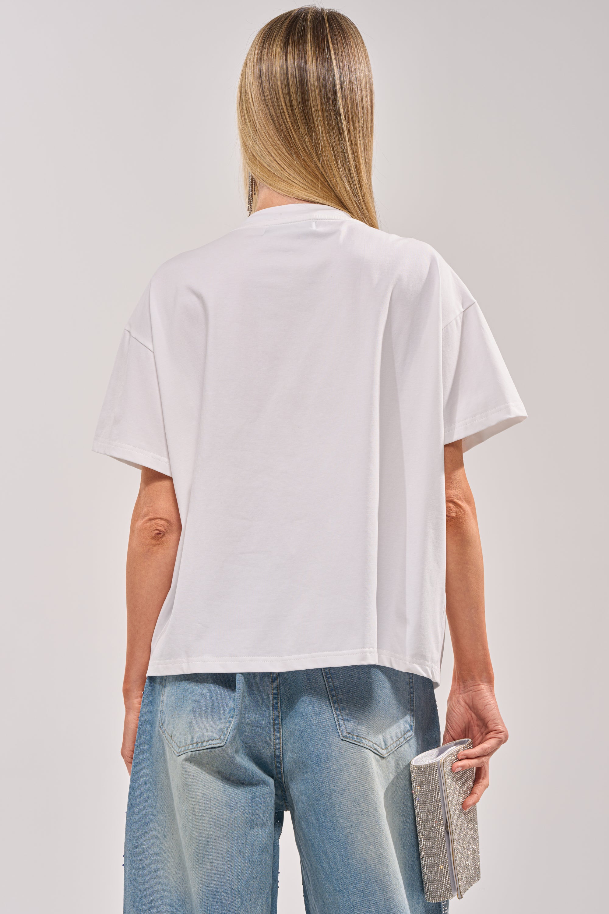 A person with long, straight blonde hair is seen from behind wearing the DIAMONDS DANCING RHINESTONE DETAIL TEE and light blue jeans, holding a sparkly rectangular clutch in their right hand. 6